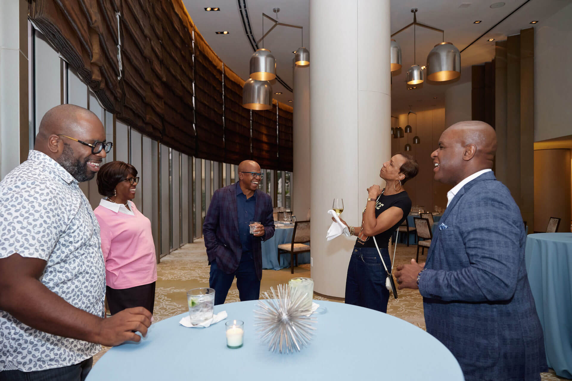 A small group chatting near tall tables in a hotel lounge, with a woman in a pink sweater and a man wearing a plaid blazer enjoying drinks, while another woman in a black T-shirt laughs with a glass of wine in hand.