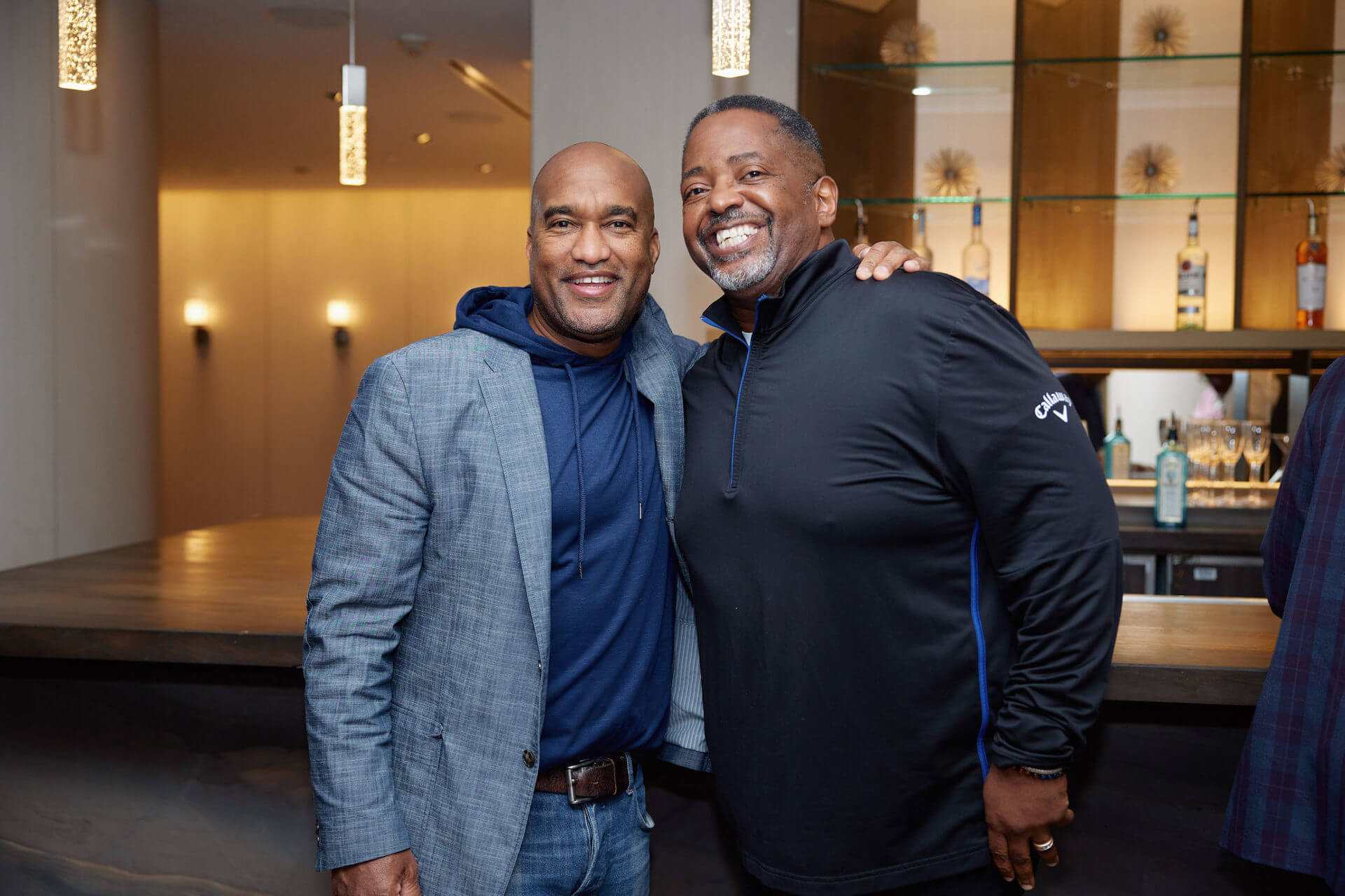 Two men standing shoulder to shoulder, one in a gray blazer over a navy hoodie, the other in a black pullover, both smiling and posing for the camera near a bar area.