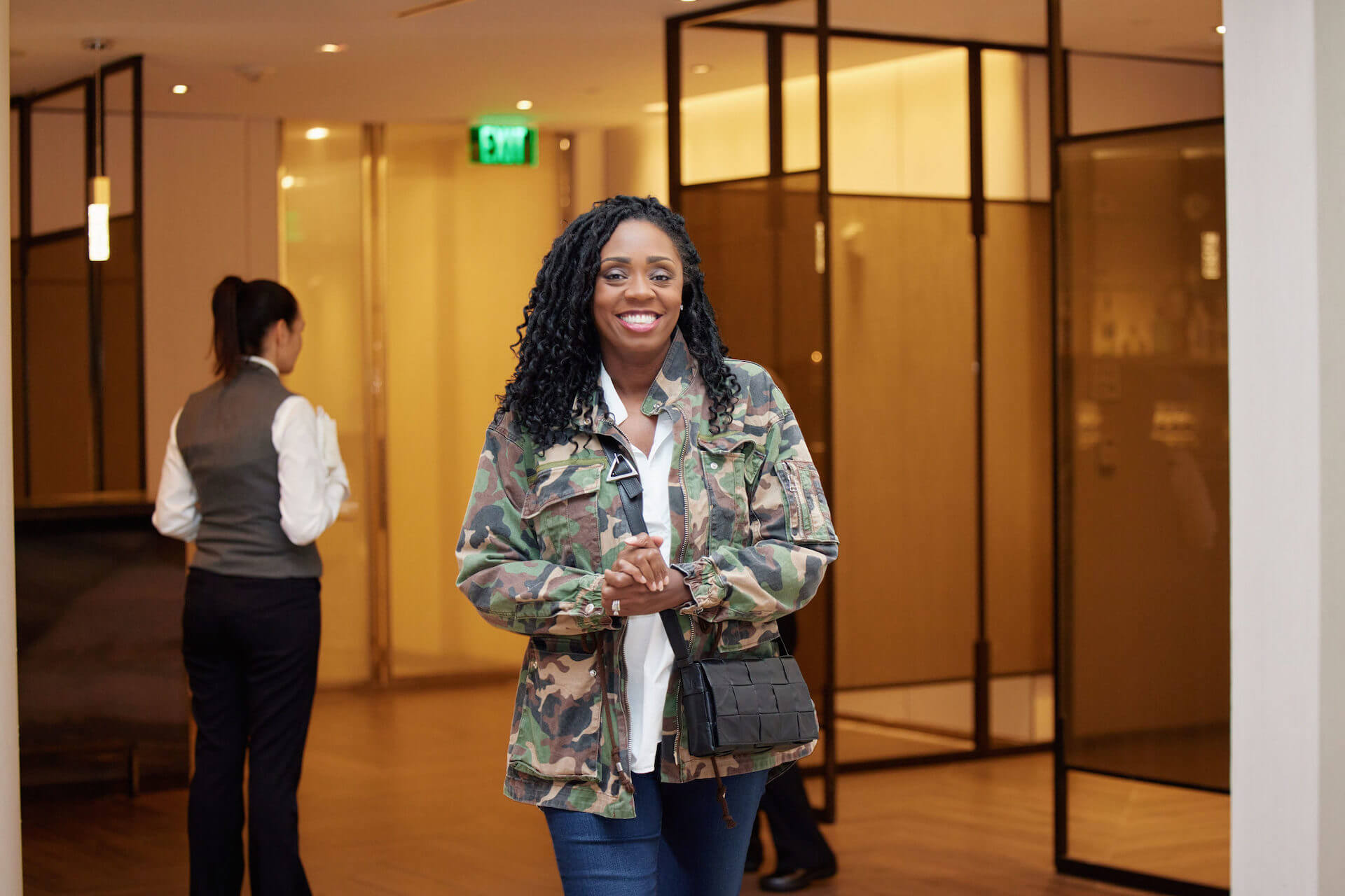 A smiling woman in a camouflage jacket walks through a lobby, hands clasped in front of her.