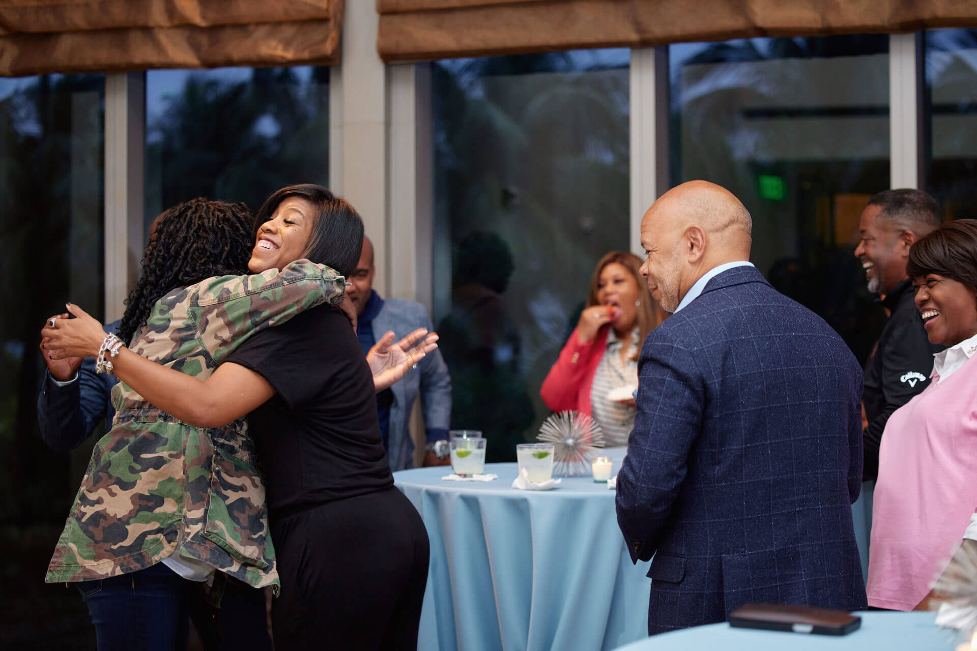 Two women share a warm hug during a gathering, surrounded by other attendees chatting and enjoying drinks.