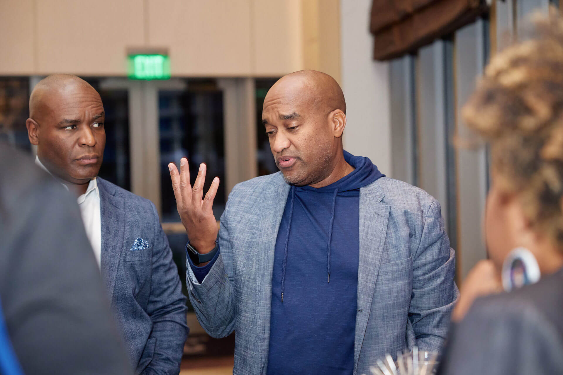 A man wearing a blazer over a hoodie gestures while speaking to another man in a suit, who listens intently.