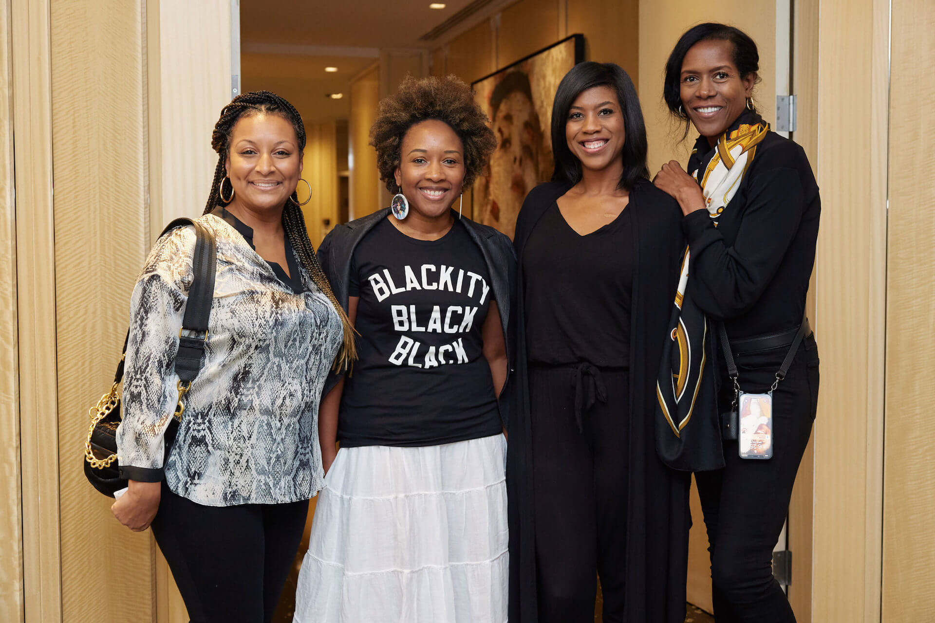 Four women stand together in a hallway, smiling at the camera. One wears a T-shirt reading “Blackity Black Black,” another wears a snakeskin-print top, and the others are dressed in black.