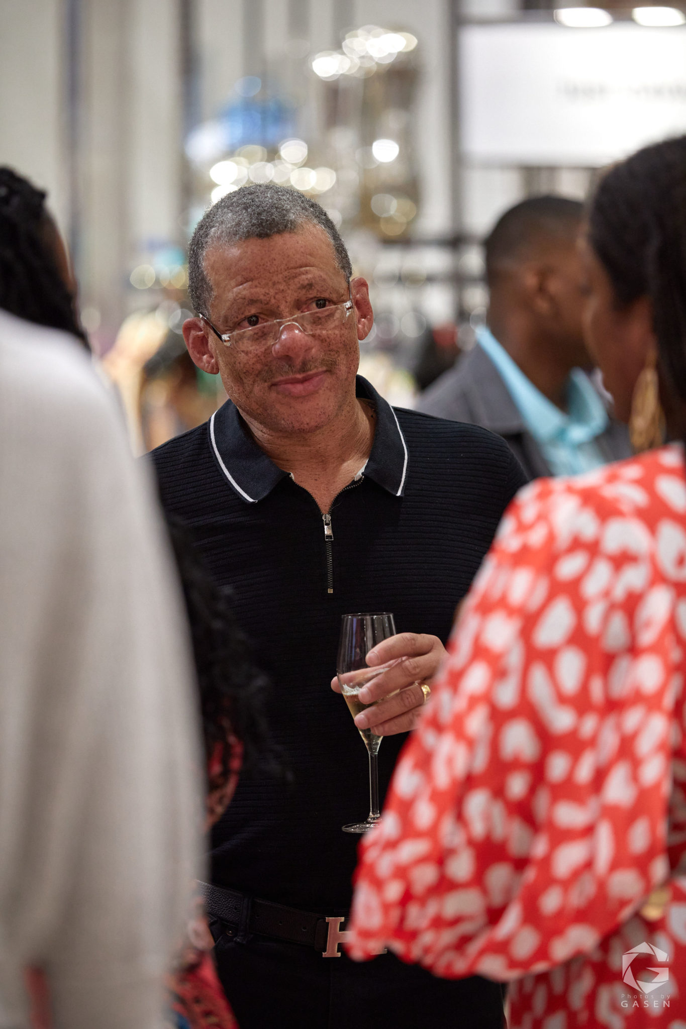 A man wearing glasses and a black polo shirt holding a glass of champagne, engaged in conversation during a retail event.
