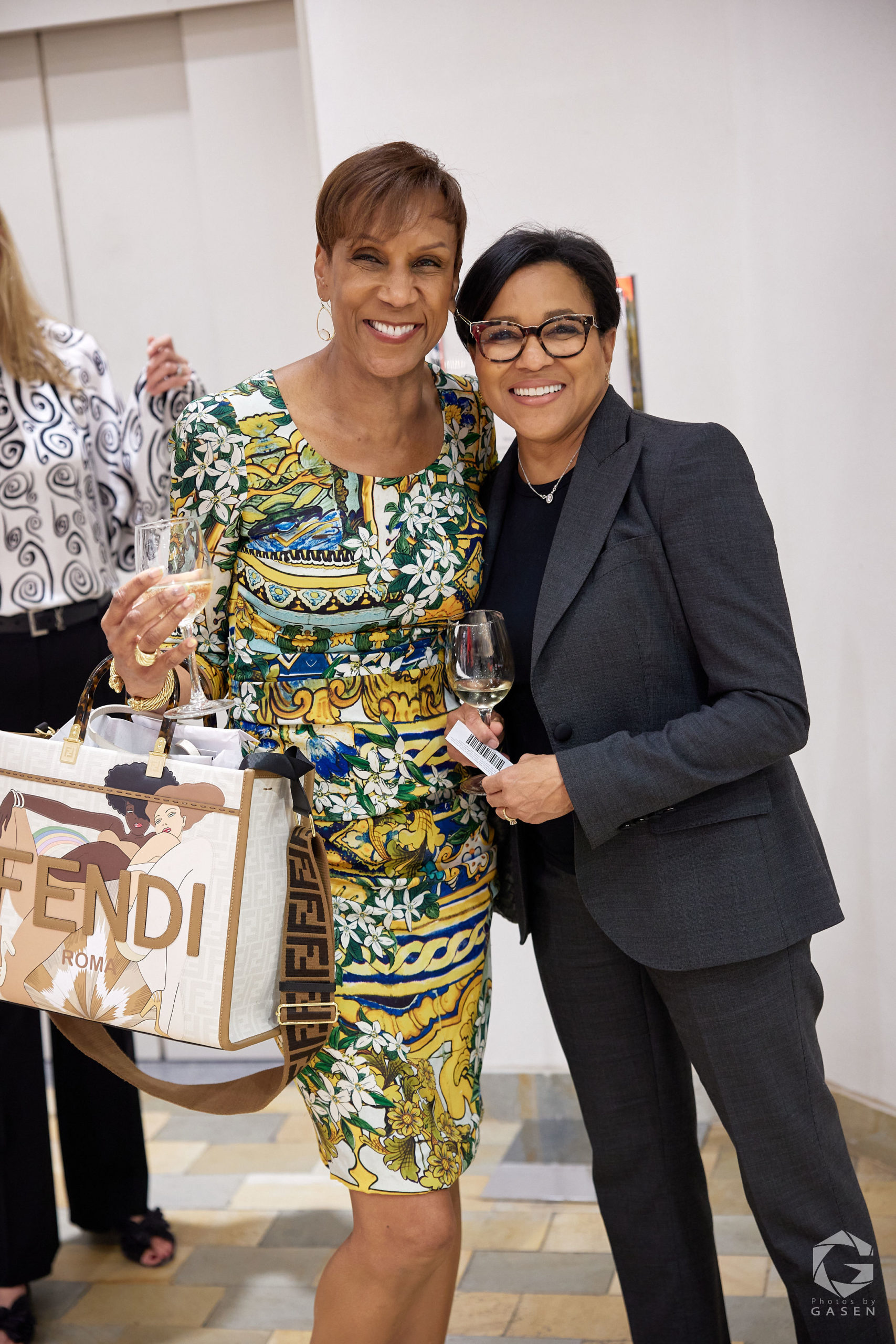 Two women smiling together. One wears a colorful floral dress and holds a designer bag, while the other is in a gray suit holding a glass of champagne.