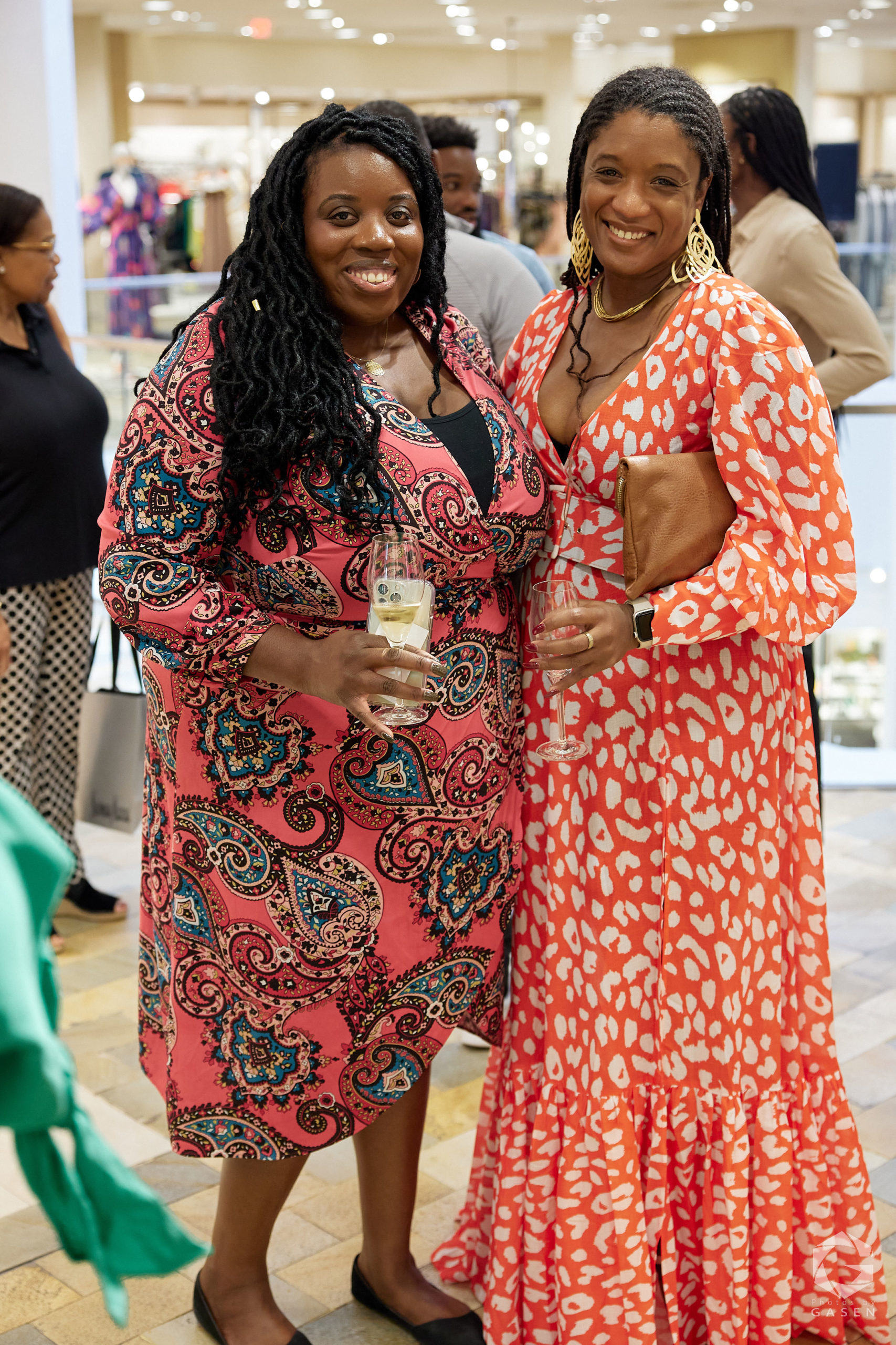 Two women standing indoors at an event, both smiling. One is wearing a pink paisley-patterned dress, and the other is in an orange dress with white spots, holding glasses of champagne.