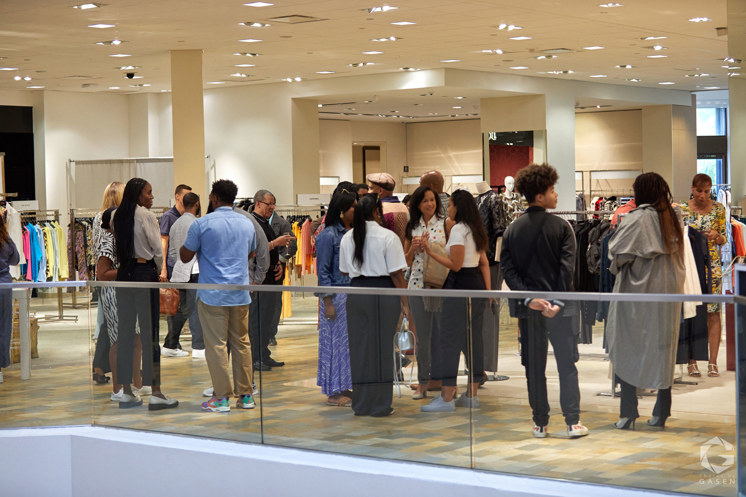 A wide shot of a retail space with groups of people mingling and chatting. Clothing racks and store displays can be seen in the background.