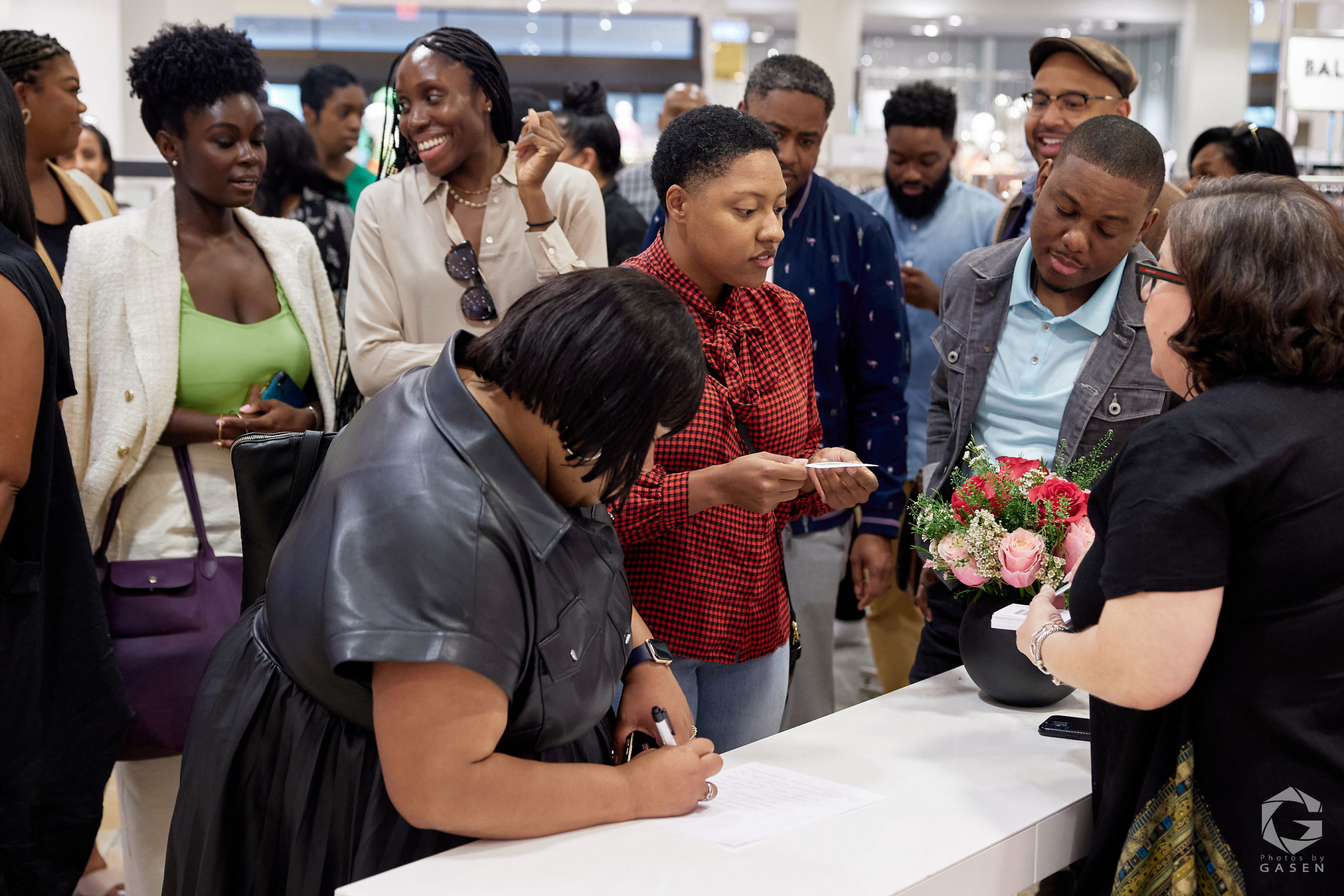 A group of people gathered around a table where a woman in black is writing. Other attendees are smiling or observing, with flowers and paperwork on the table.