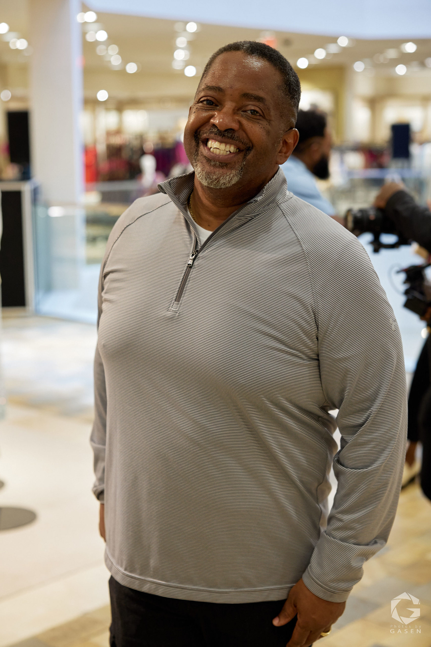 A man in a light gray pullover smiles brightly at the camera in a retail store setting.