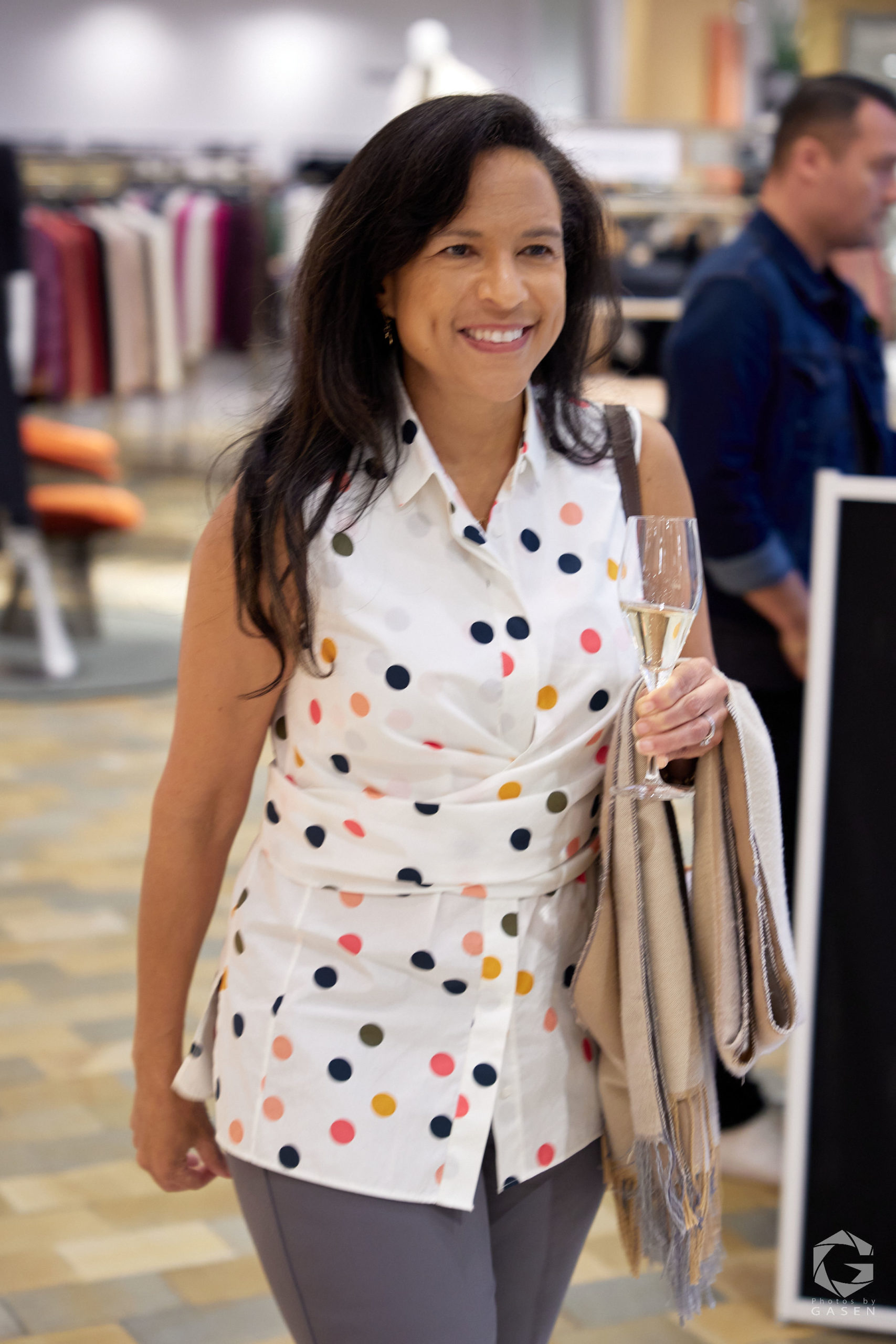 A woman in a white polka dot top and gray pants holds a glass of champagne and a scarf, smiling as she walks in a retail space.