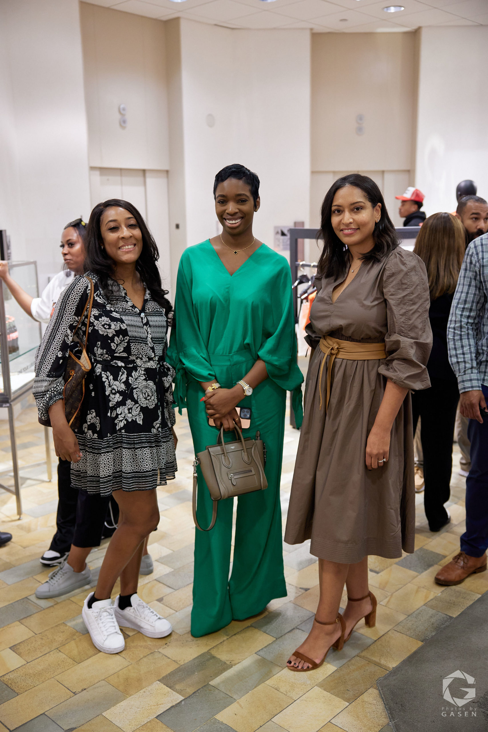 Three women standing and smiling at an event. One wears a black floral dress, another is in a green jumpsuit, and the third is in a brown dress. They are posing indoors in a retail space.