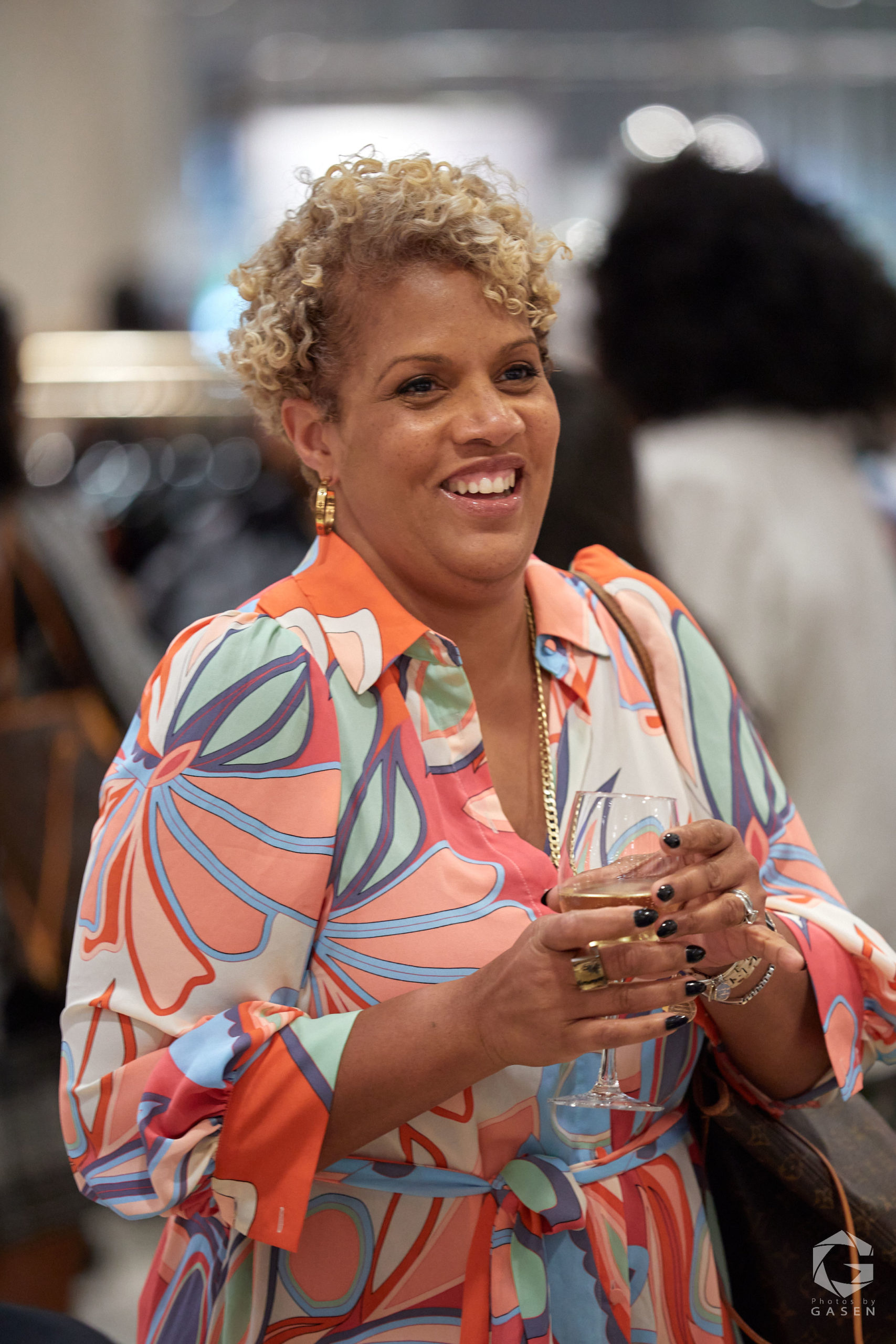 A woman with short curly blonde hair, wearing a colorful patterned blouse, smiles brightly while holding a glass of champagne.