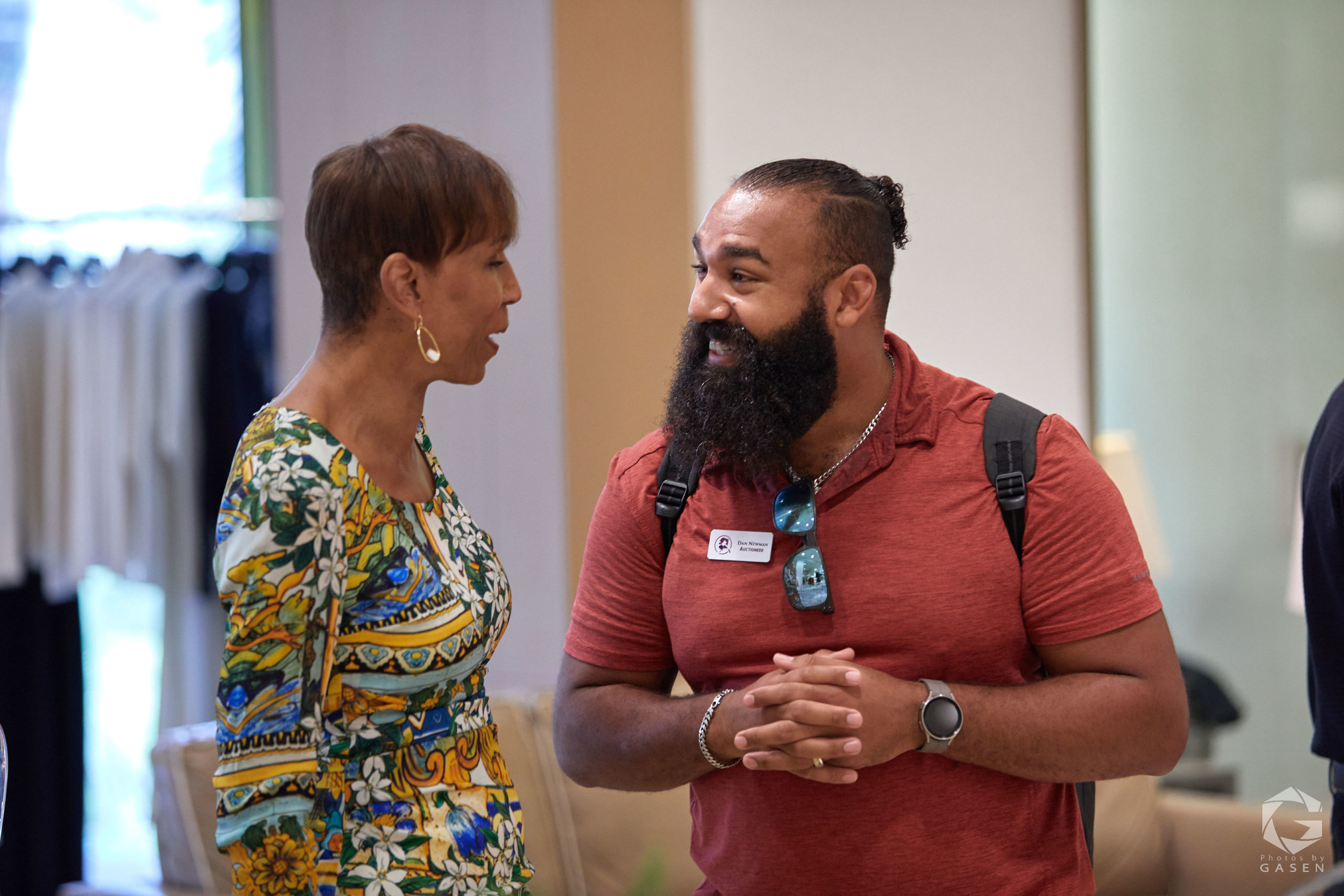 A man with a beard and red shirt chats with a woman in a colorful patterned dress. They are smiling and standing in what appears to be a store or event space.