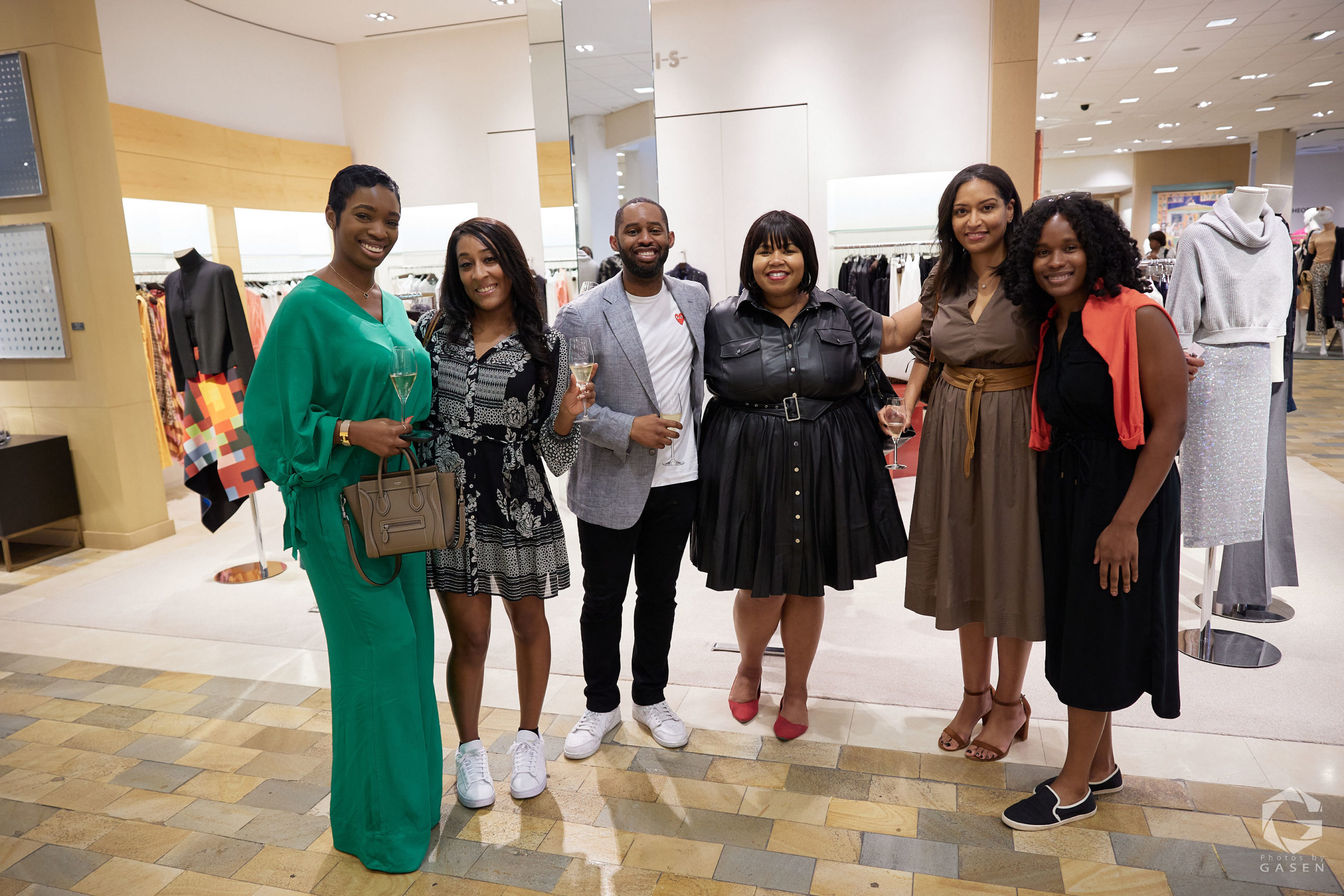 A group of six people standing in a retail space, all smiling. The group includes men and women dressed in colorful and stylish outfits, holding glasses of champagne.