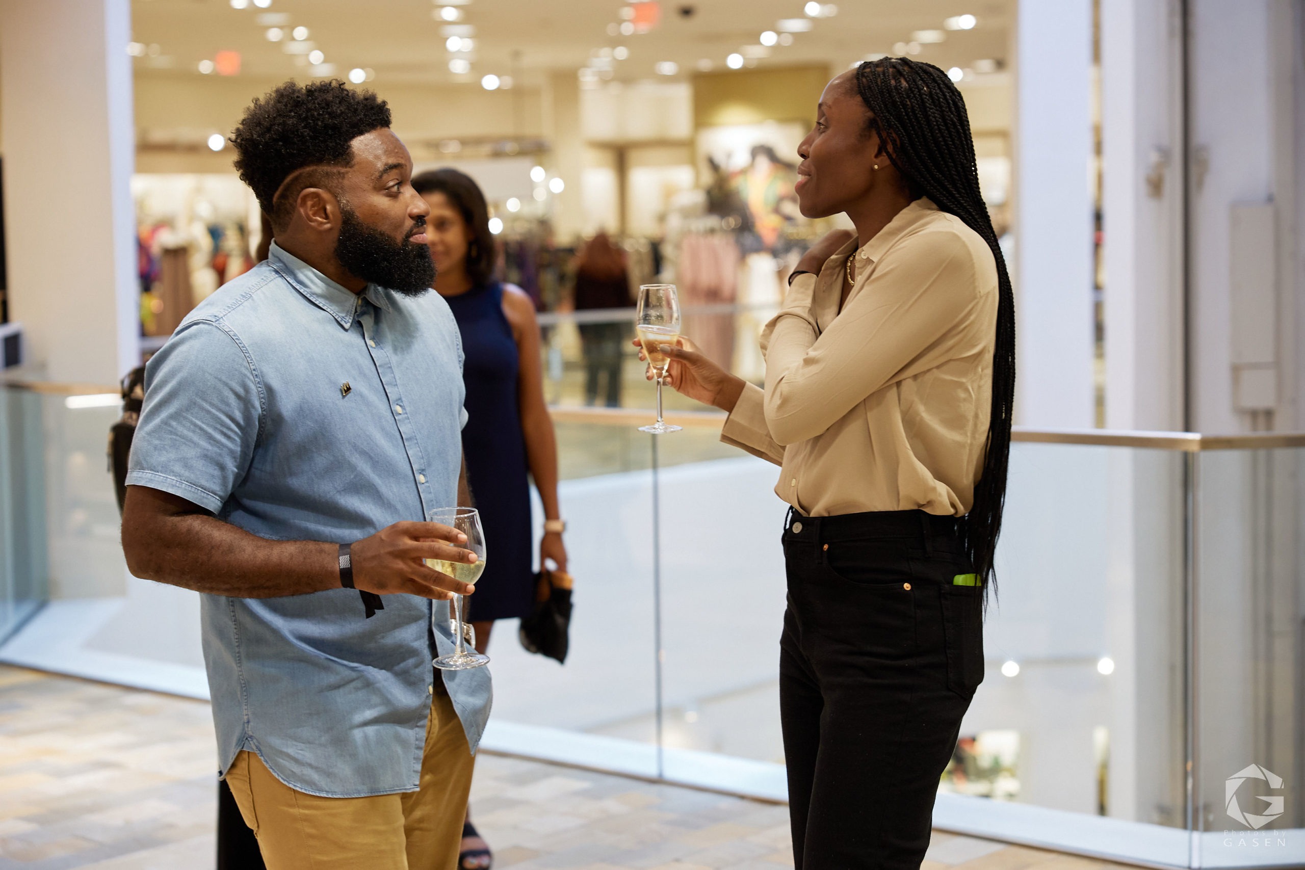 A man in a denim shirt and a woman in a beige blouse with braids share a conversation while holding glasses of champagne at an indoor event.