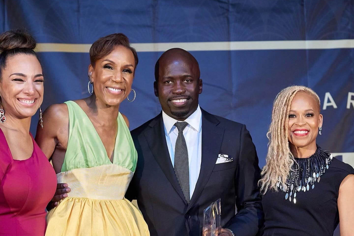 Four elegantly dressed individuals stand together at an awards event, smiling at the camera. The man in the center, wearing a black suit and tie, holds a glass award. The women surrounding him wear vibrant dresses, including a green and yellow gown, a magenta dress, and a black dress with decorative embellishments. They are positioned in front of a dark blue backdrop, exuding joy and celebration.
