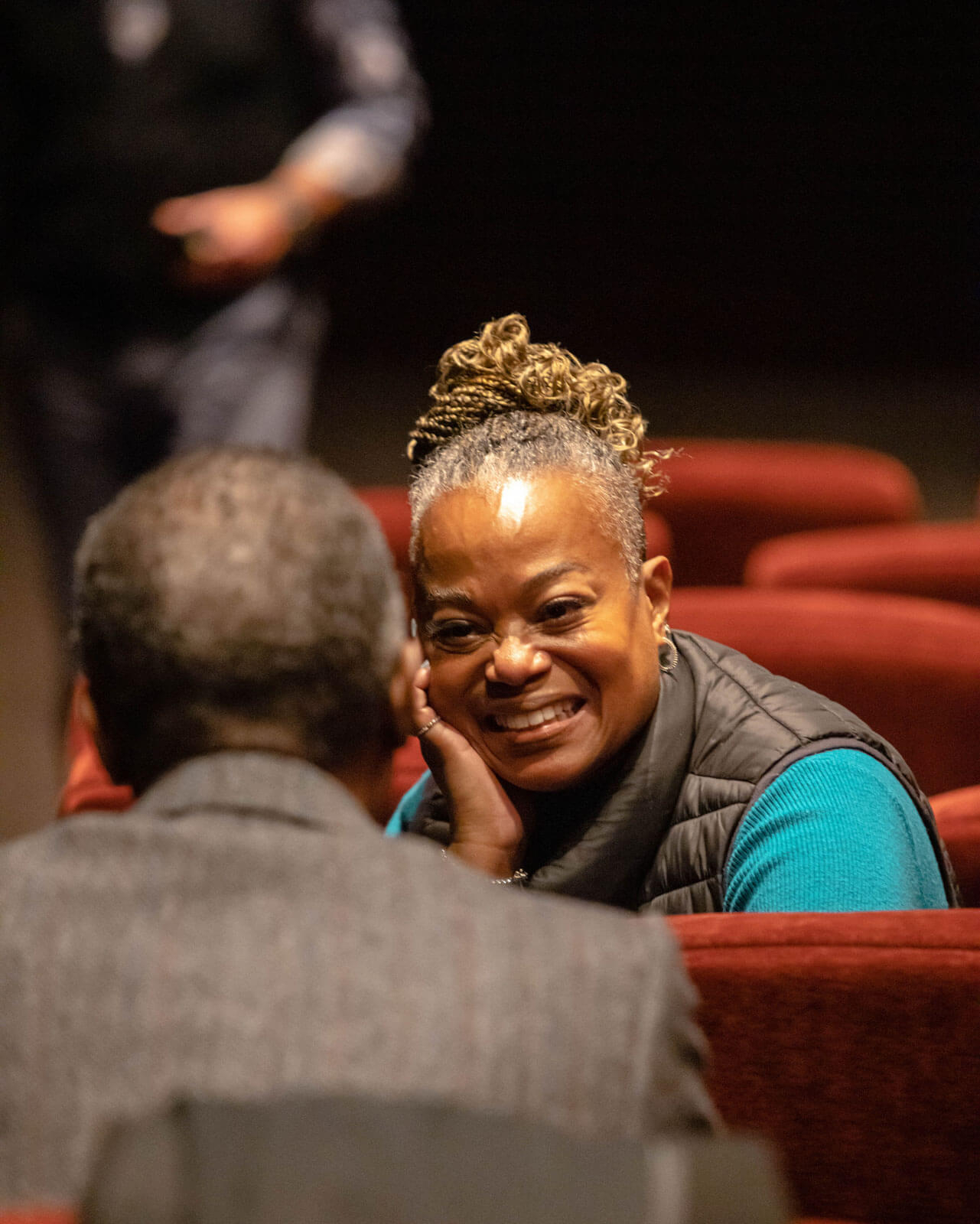 A woman with braided hair smiles warmly while engaged in conversation with someone seated across from her in a theater setting. She is wearing a black vest over a blue sweater and leans on her hand in a relaxed posture. The background features red theater seats, and a blurred figure is visible in the distance, adding to the intimate and engaging atmosphere of the moment.