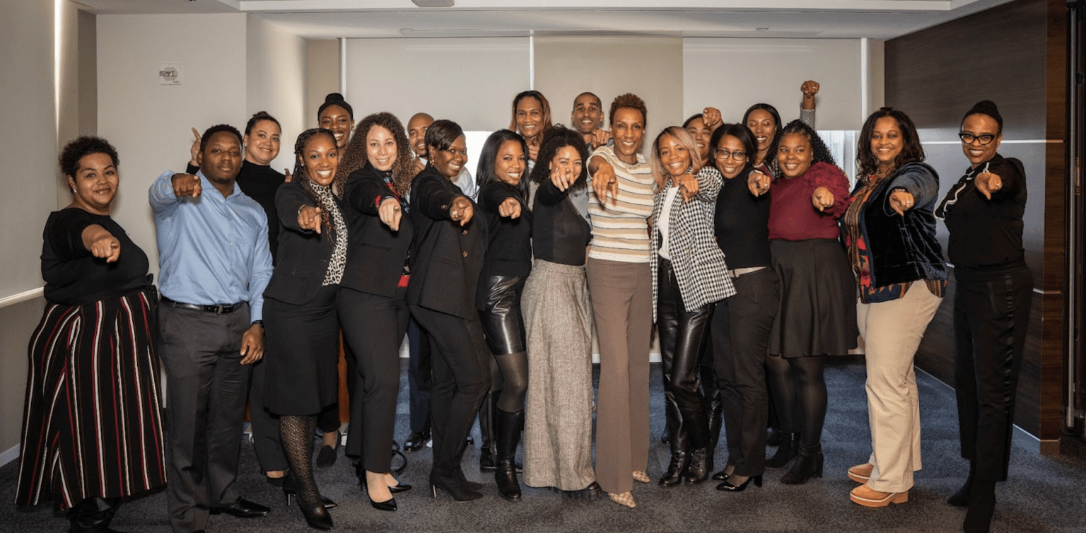 A diverse group of professionals posing together, smiling, and pointing toward the camera in a well-lit office space.