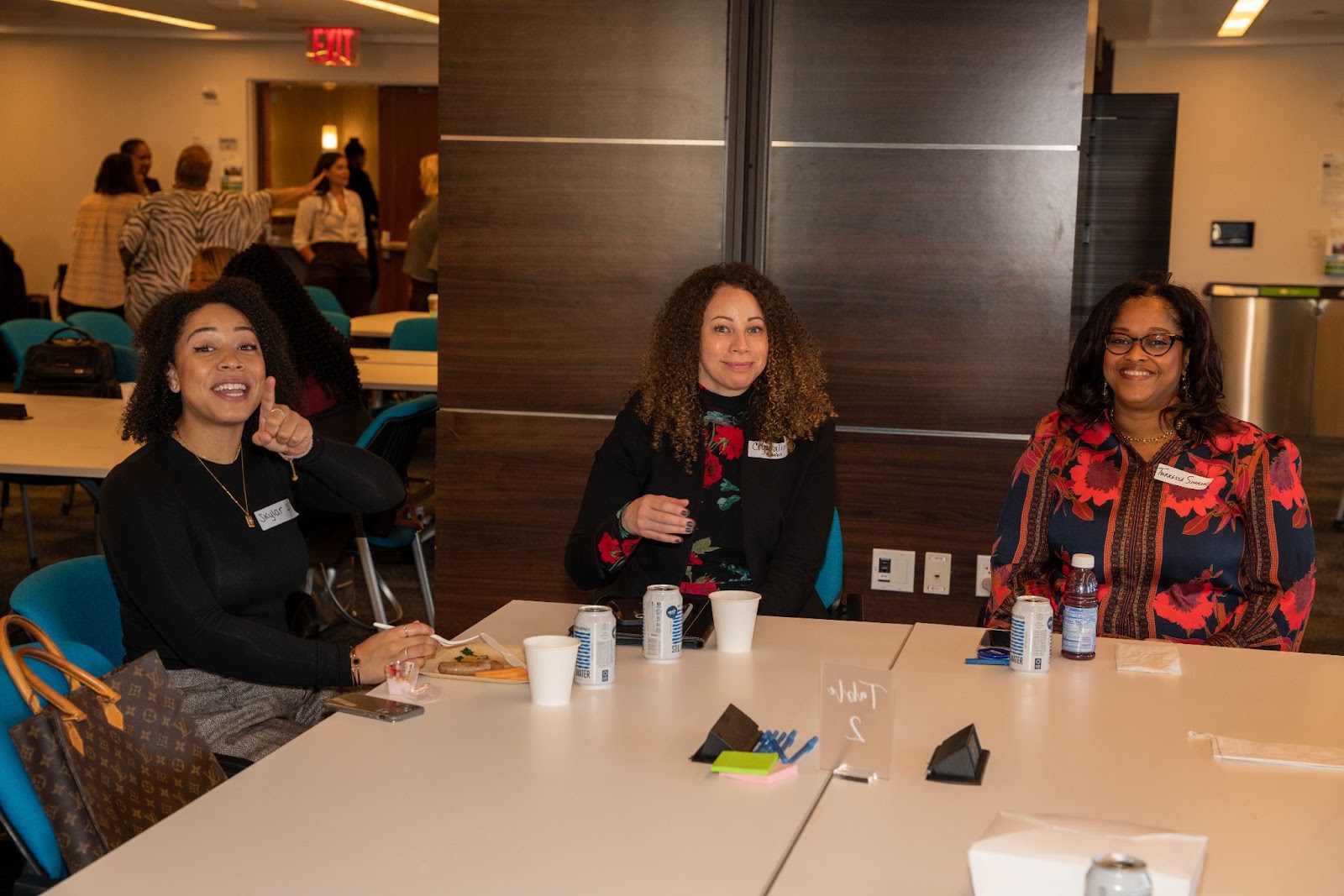 Three women sit together at a conference table, smiling and talking while enjoying snacks and drinks.