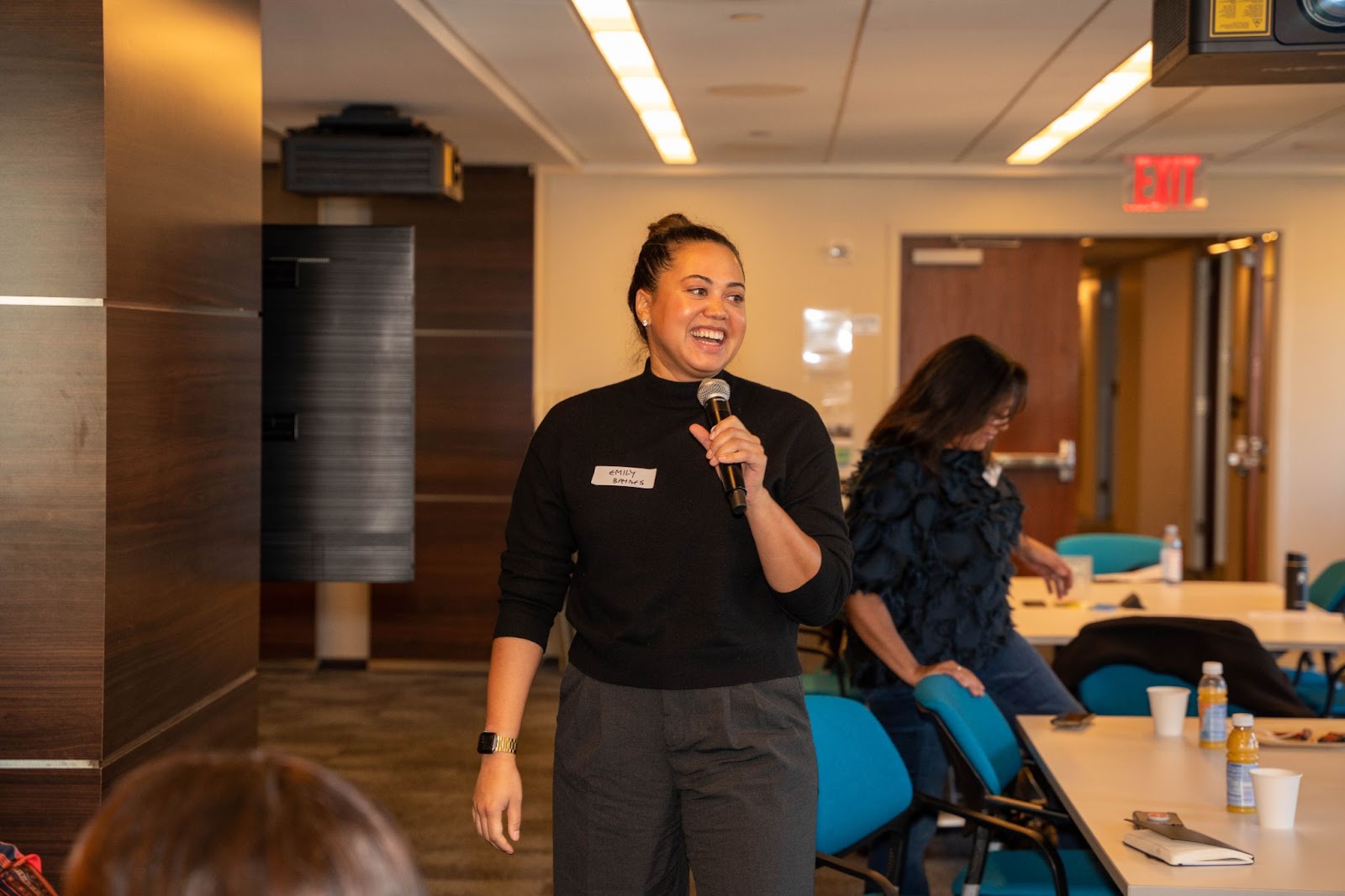 A woman in a black sweater laughs warmly as she speaks into a microphone during an event.