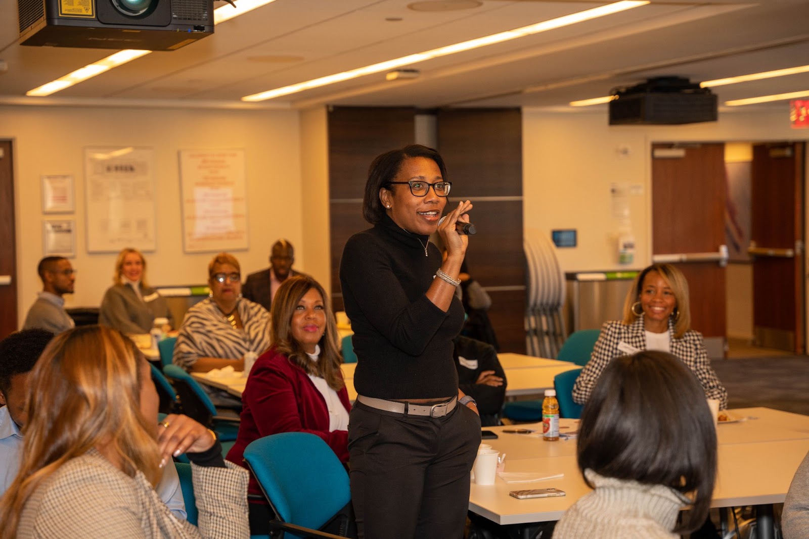 A woman speaking into a microphone while standing in front of an audience in a professional workshop setting.