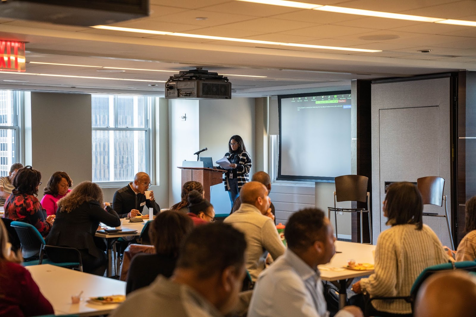 A conference room with attendees seated at tables, focusing on a speaker presenting at a podium in front of a screen.