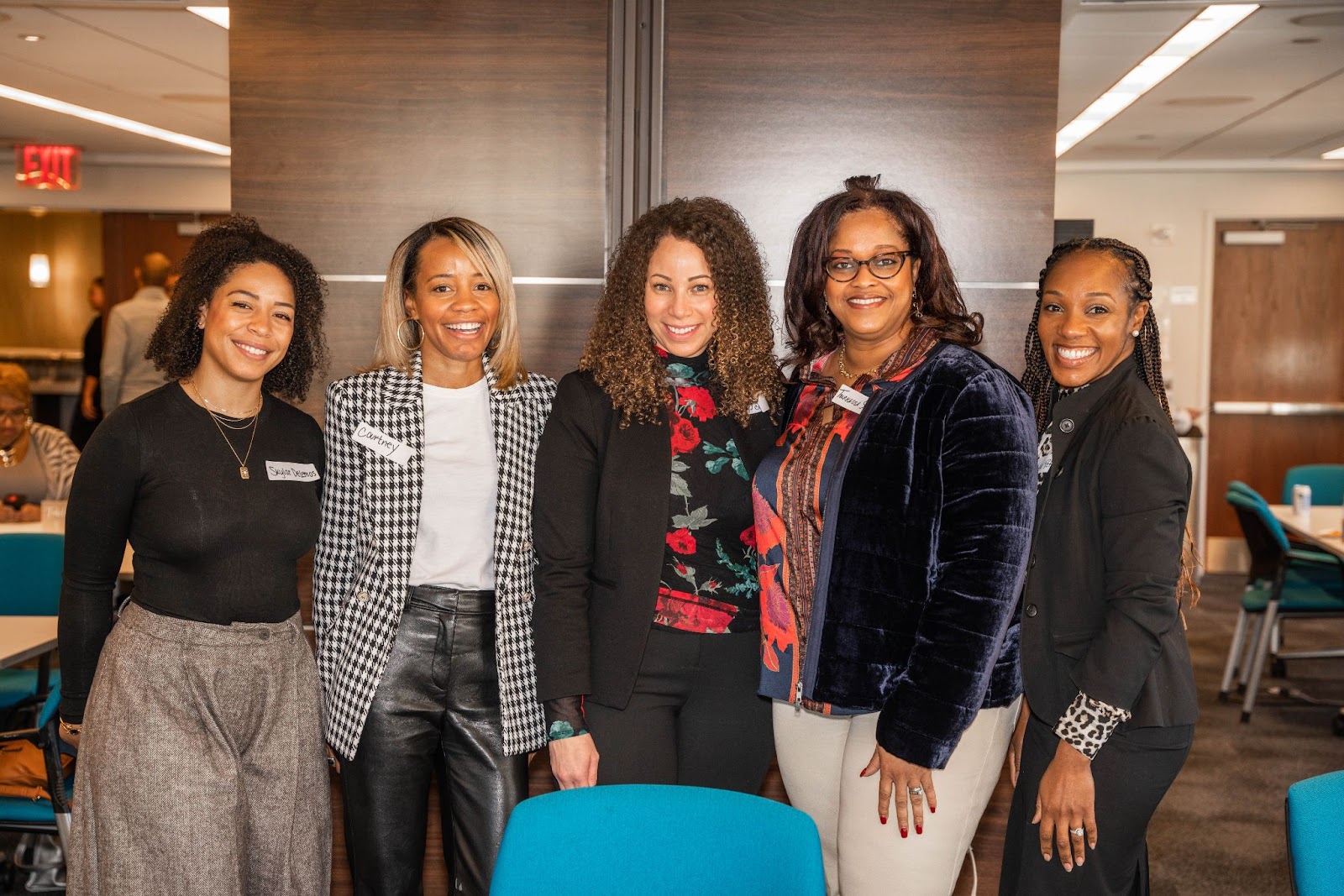 A group of five women posing together, smiling, with name tags visible. They are standing in a professional setting with a modern interior.