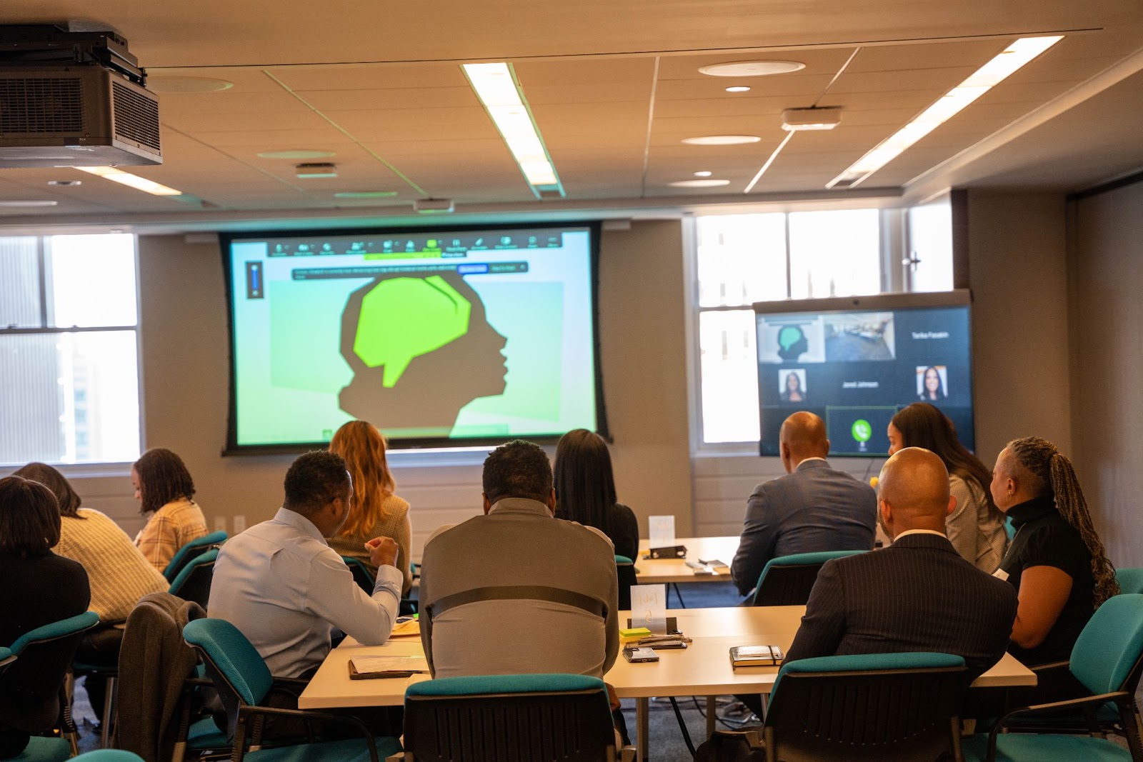 Attendees seated at tables, attentively watching a presentation displayed on a large screen, showing an illustration of a green brain silhouette.