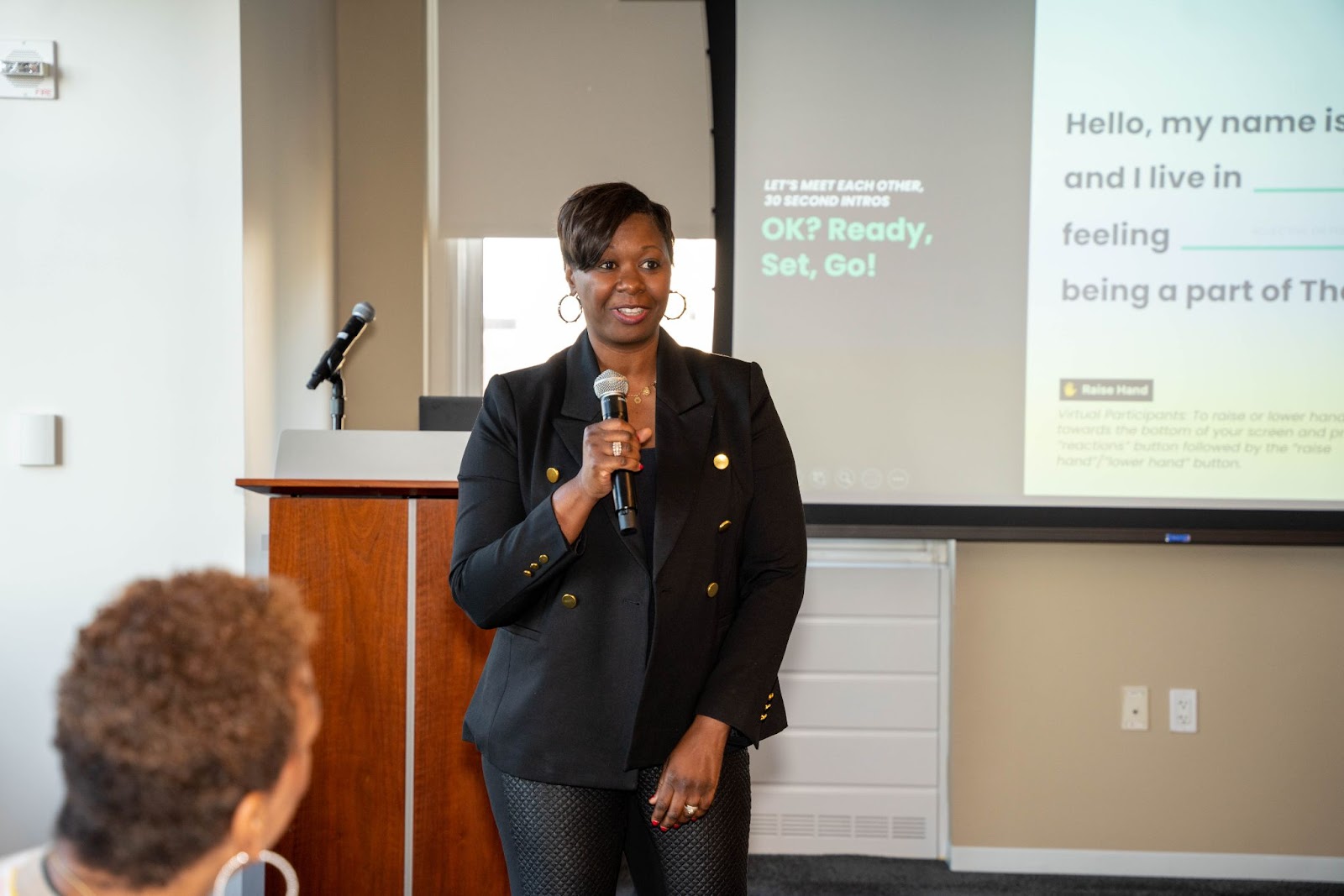 A woman in a black blazer speaks into a microphone in front of a projected slide that encourages introductions.