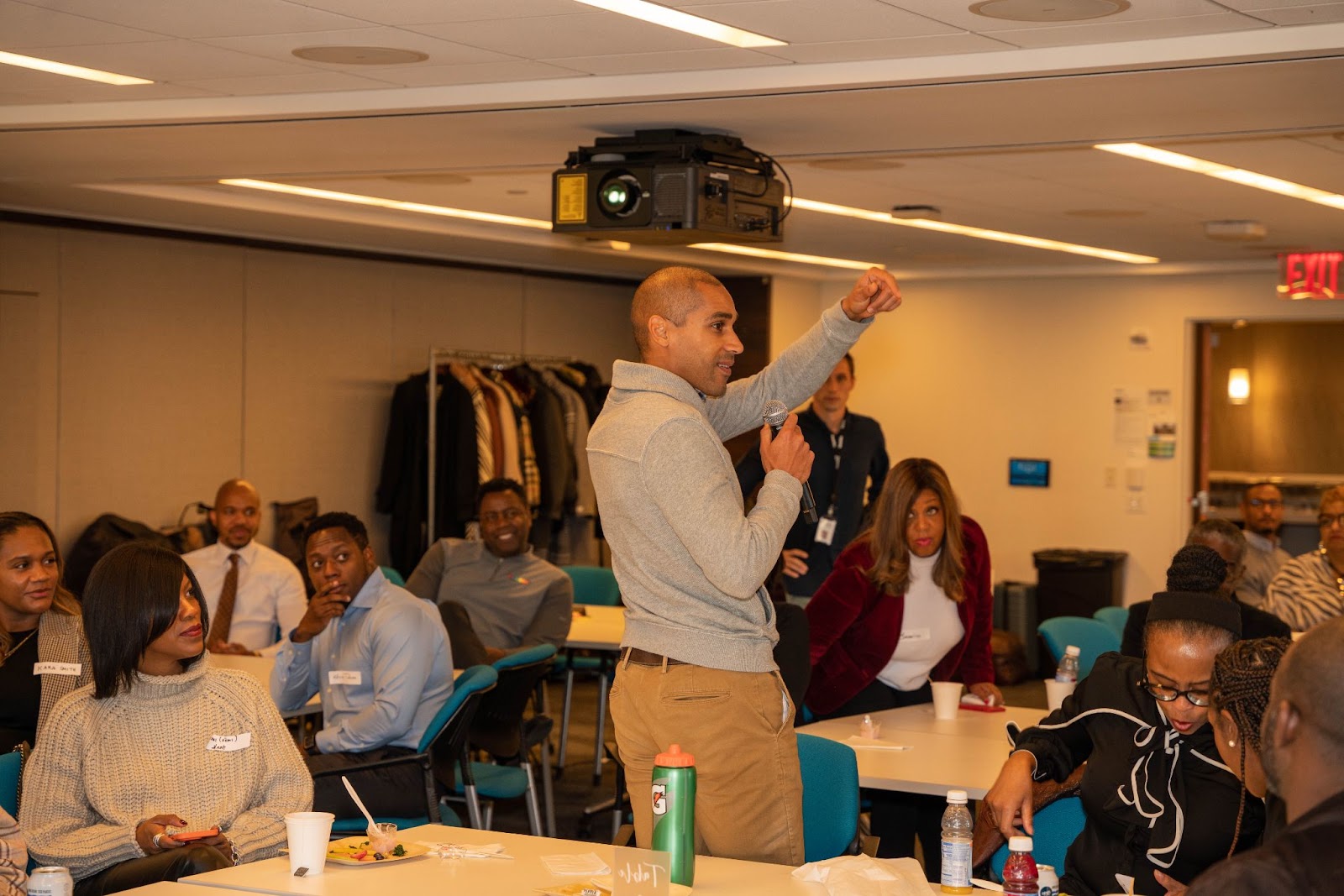 A man in a gray sweater stands and speaks passionately into a microphone, gesturing as others around the room look on attentively.