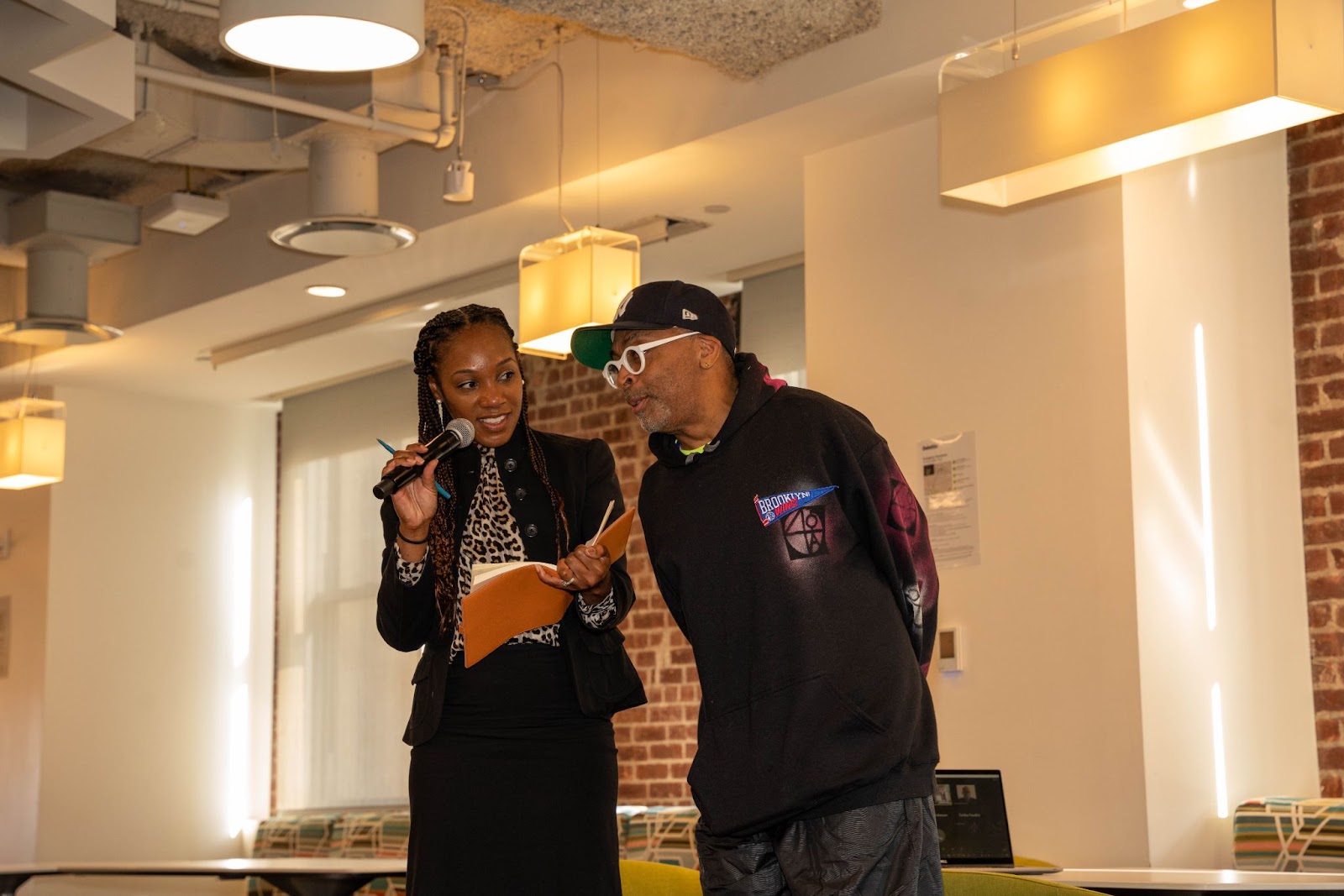 A young woman holding a microphone and a notebook stands next to a man in a hoodie and hat during a lively conversation in a bright, modern space.
