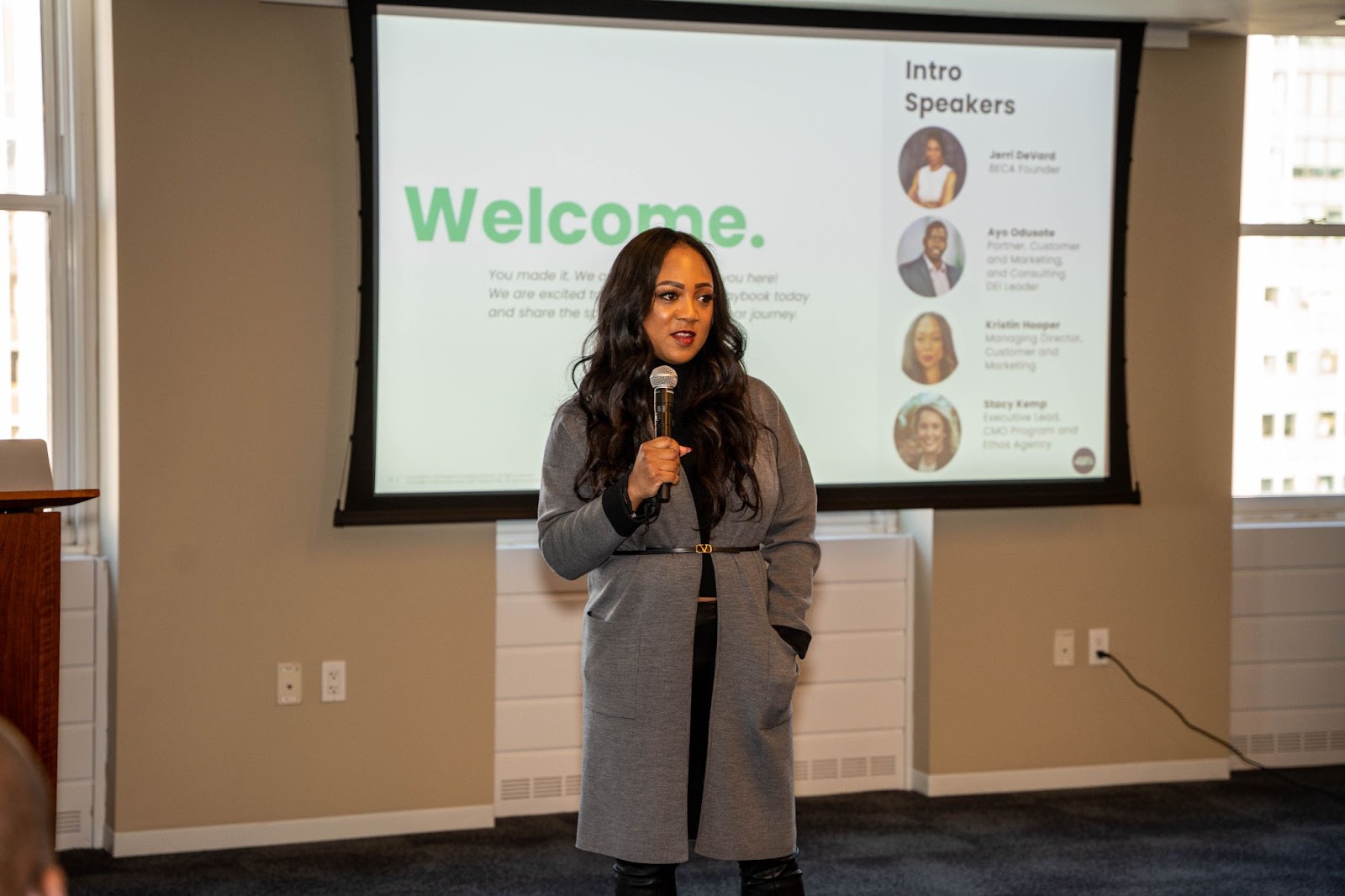 A woman in a gray coat speaks into a microphone during a presentation, with a "Welcome" slide projected in the background.