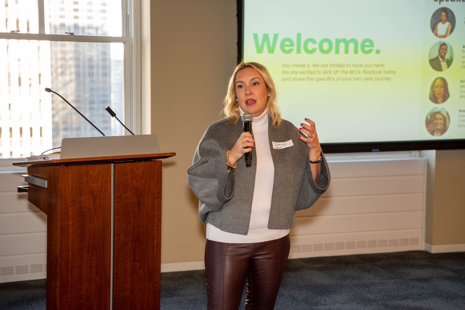 A woman in a gray cape-style jacket speaks into a microphone while presenting in front of a projector screen displaying a "Welcome" slide.