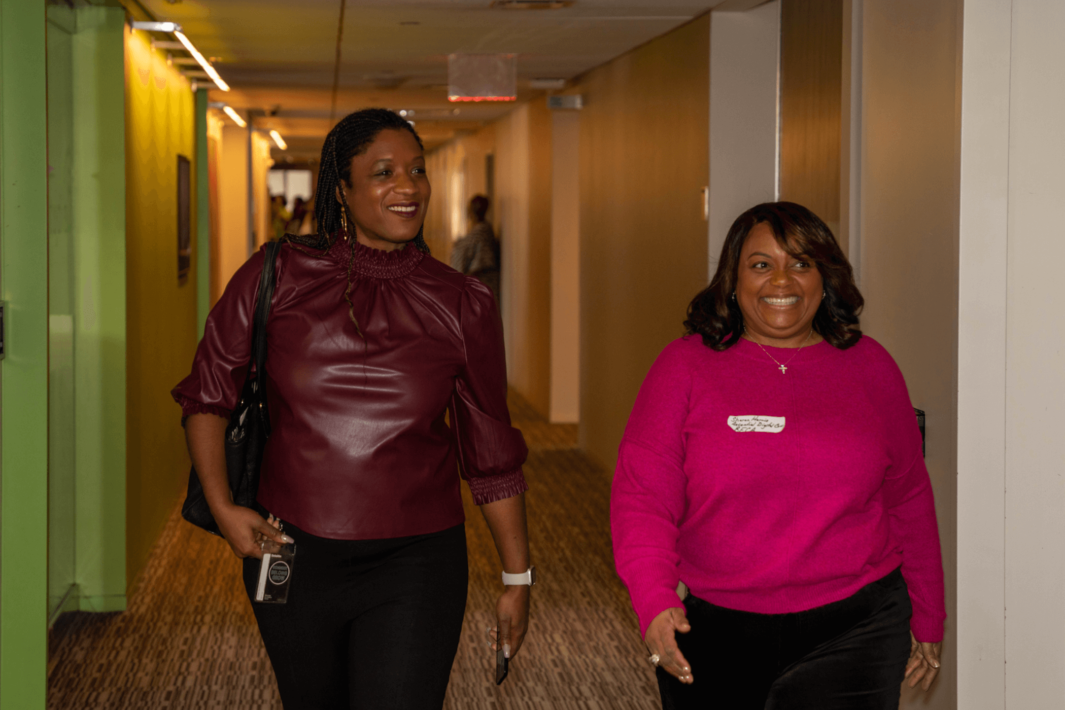 Two women walking down a hallway, one wearing a burgundy top and the other in a bright pink sweater, smiling and chatting.