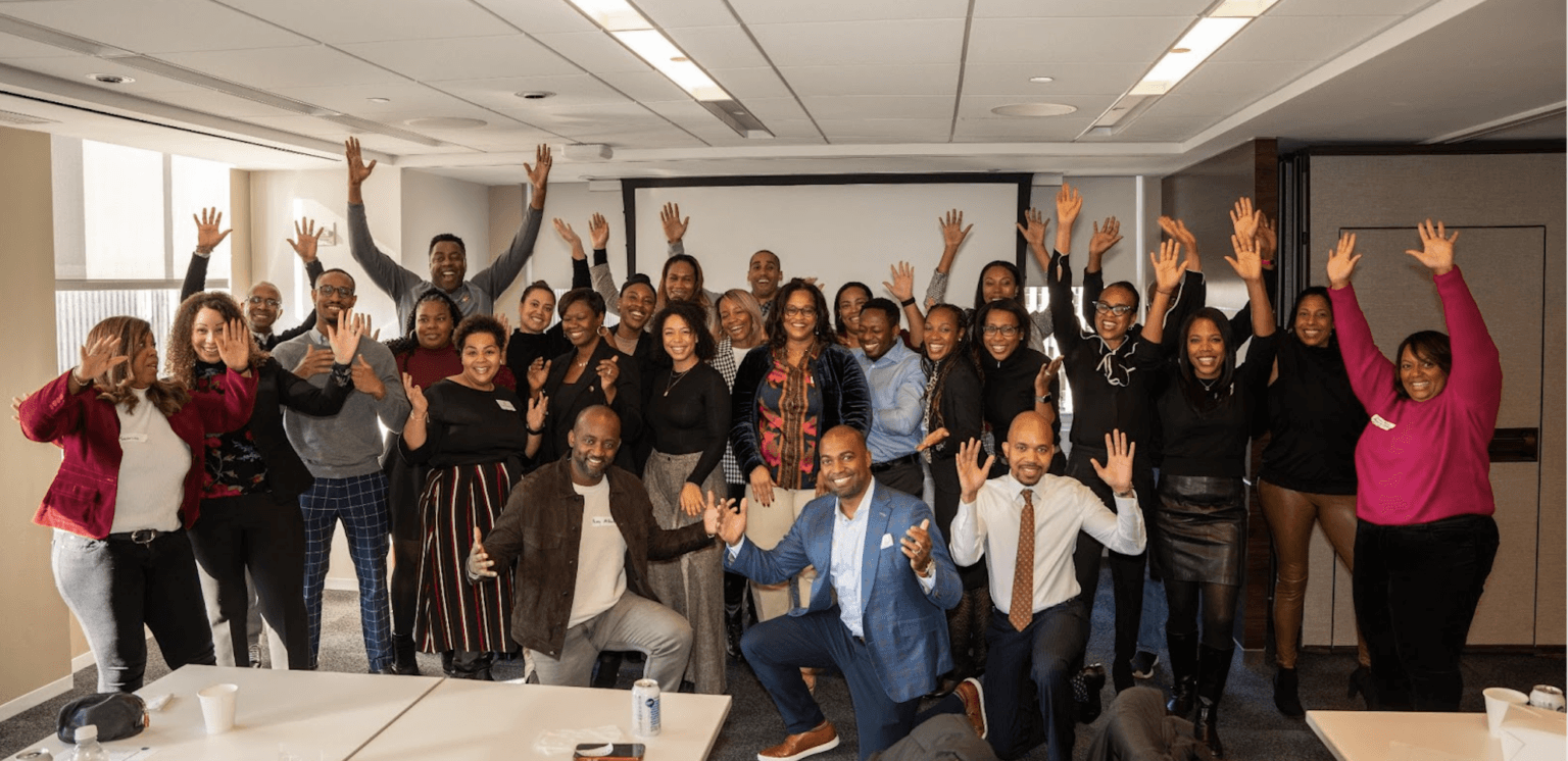 A large group of people gathered in a bright meeting room, posing energetically with their hands raised, showcasing a joyful and collaborative atmosphere.