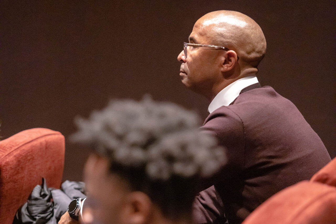 A bald man wearing glasses and a dark brown sweater over a white collared shirt sits attentively in a theater setting. His focused expression suggests deep engagement in the event. The blurred foreground shows the back of another attendee’s head, while red theater seats and a dimly lit background add to the ambiance of a professional or formal gathering.