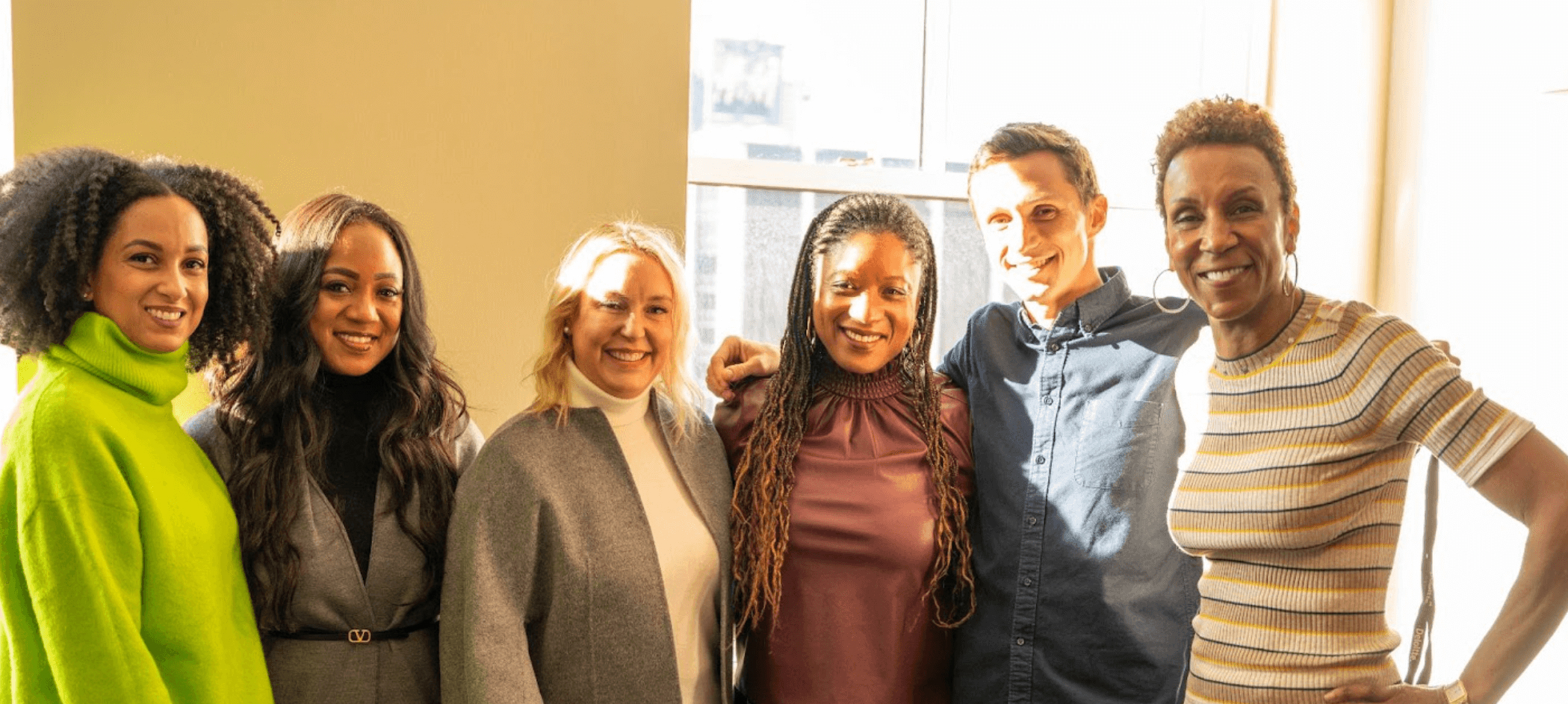 A group of six people, diverse in age, gender, and ethnicity, stand together in a warmly lit indoor space, smiling at the camera. They are dressed in stylish yet professional attire, with one individual wearing a vibrant green sweater. Sunlight streams through a large window behind them, creating a bright and welcoming atmosphere.