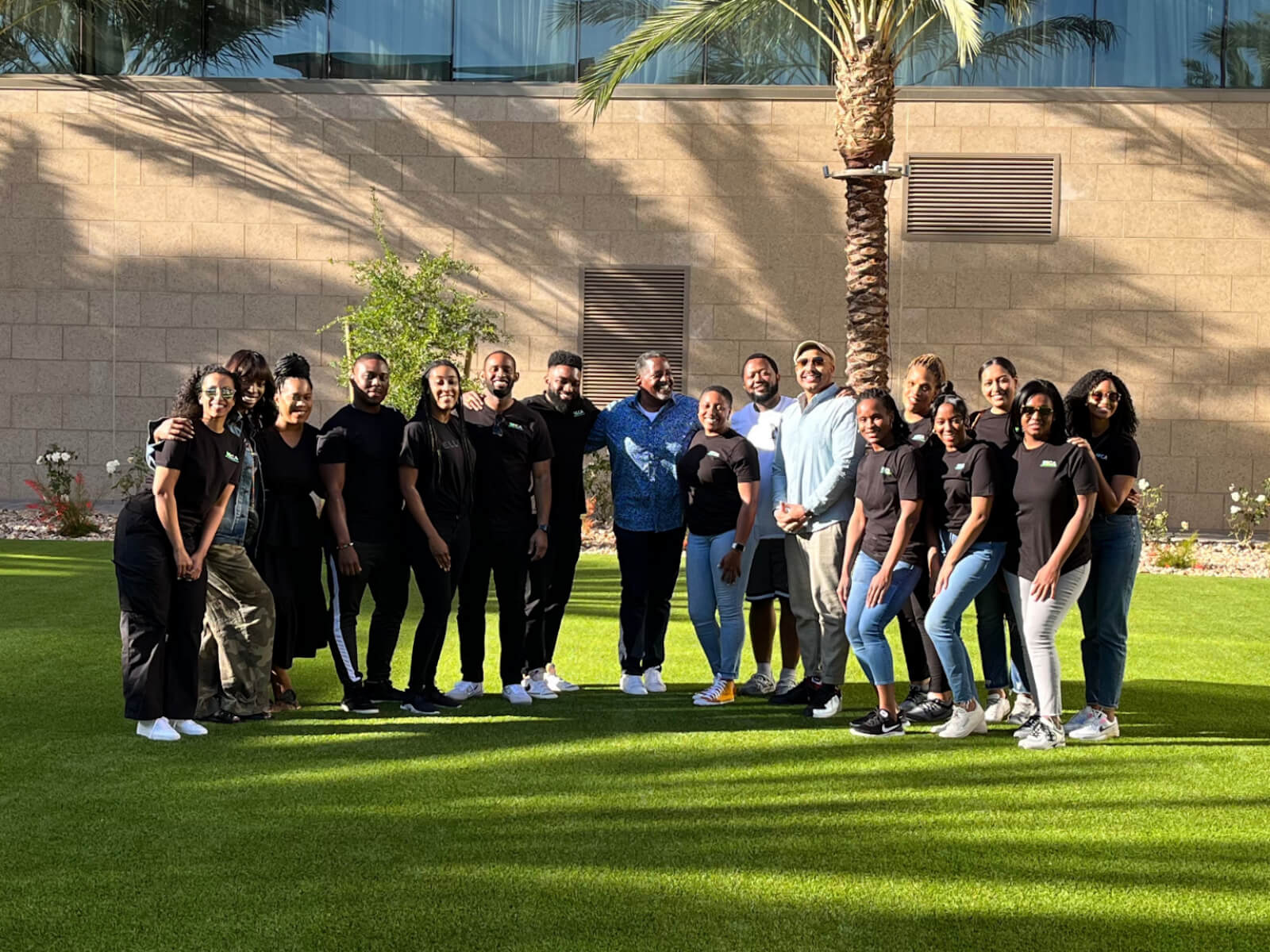 A large group of people posing together outdoors on a lush green lawn, with a building and palm trees in the background, showcasing camaraderie and teamwork.