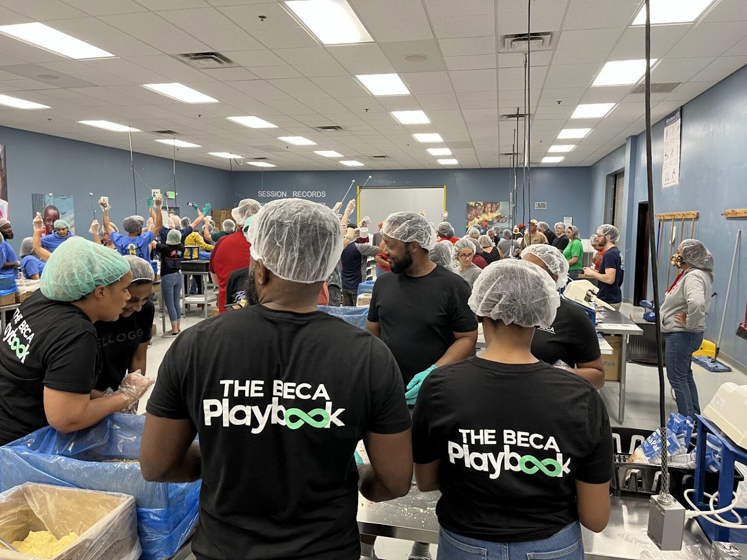 A room full of volunteers in hairnets, wearing black T-shirts with “The BECA Playbook,” busily packing items at a large food packaging station.