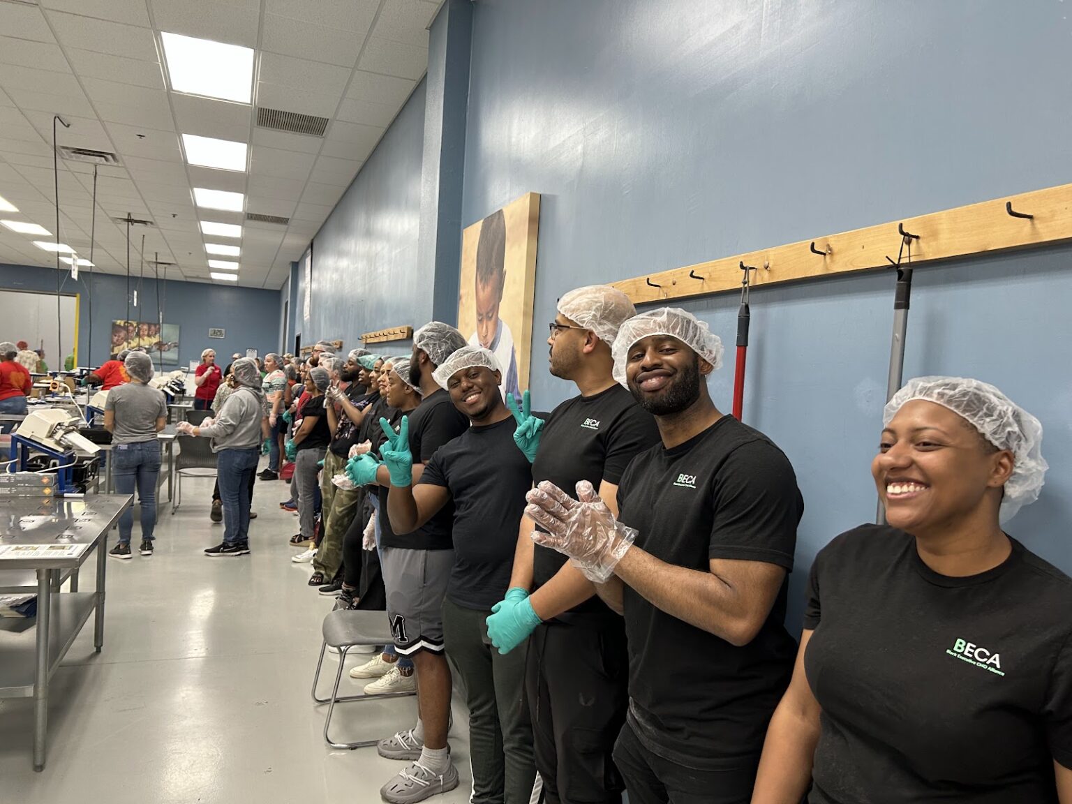 A row of smiling volunteers in black “BECA” T-shirts and hairnets, standing against a blue wall, with other teams working in the background.