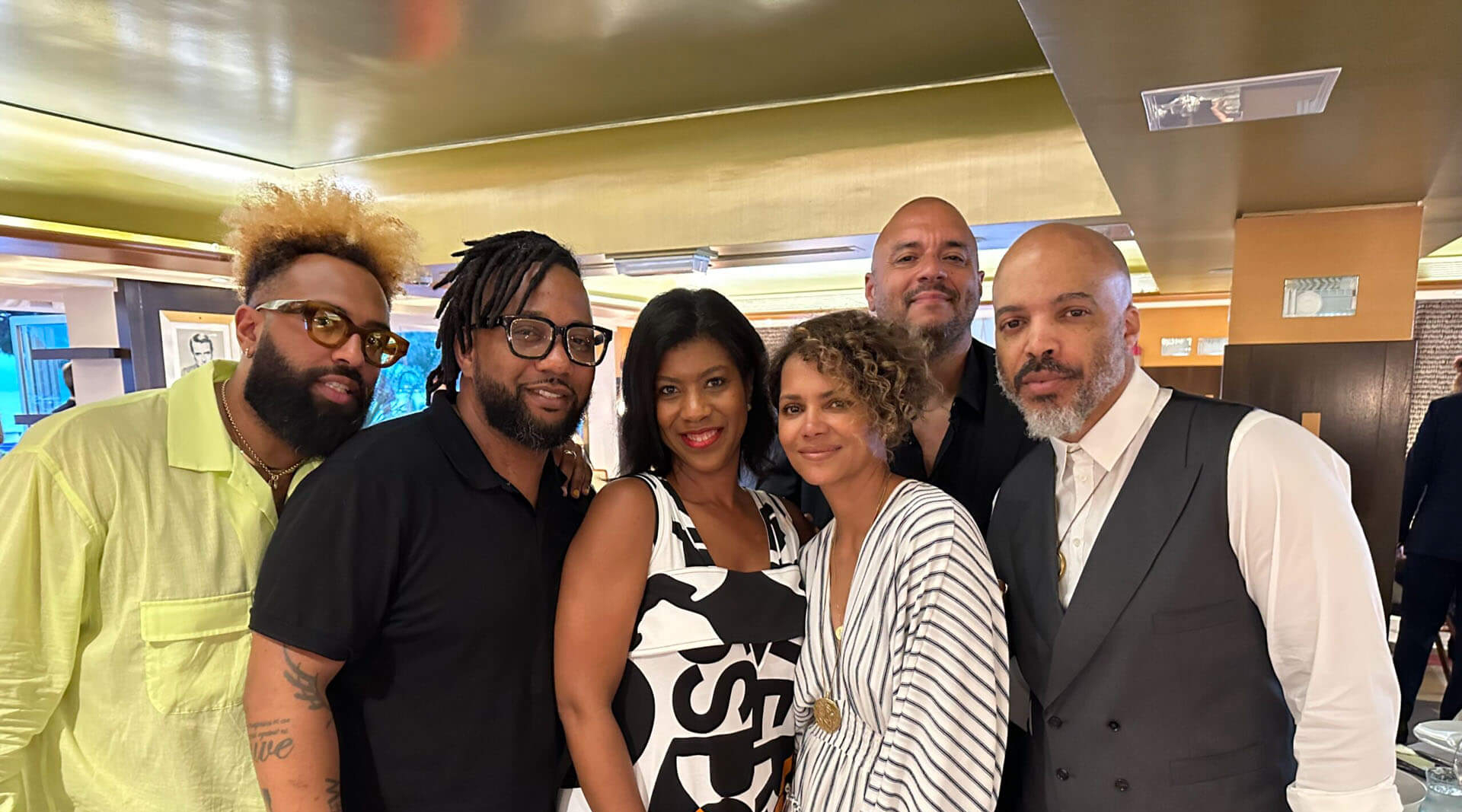 A group of six stylish individuals poses together at an indoor event, smiling warmly at the camera. They are dressed in a mix of casual and elegant attire, including patterned outfits, vests, and bold accessories. The setting features warm lighting and sophisticated decor, suggesting an upscale social or networking gathering.