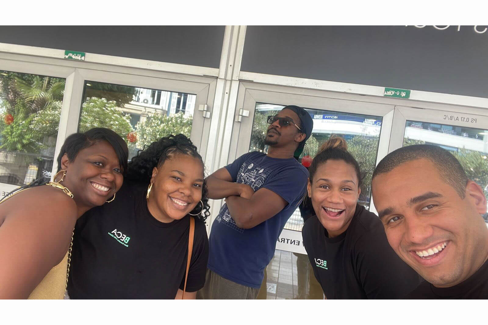 A group of five people poses together outside a building, smiling and having fun. Two individuals wear BECA branded shirts, while another strikes a playful pose in sunglasses with arms crossed. The setting includes glass doors reflecting greenery and an urban environment in the background. Their expressions and body language convey a relaxed and joyful atmosphere.