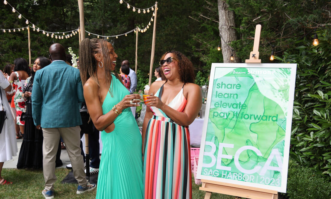 Two women laugh and clink glasses at an outdoor BECA Sag Harbor 2023 event. One wears a flowing green dress, and the other wears a colorful striped dress and sunglasses. They stand next to a sign that reads "Share, Learn, Elevate, Pay it Forward." The background features elegantly dressed guests mingling under string lights, creating a festive and sophisticated atmosphere in a lush, green setting.