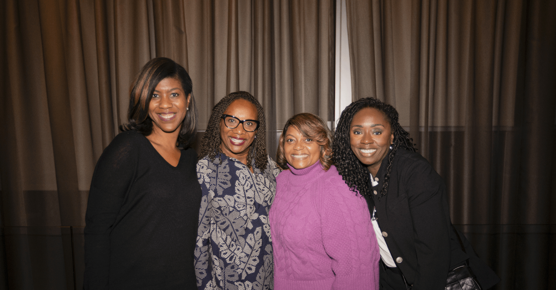 Four women stand together, smiling at the camera in an indoor setting with a backdrop of elegant drapery. They are dressed in stylish, professional attire, including a black sweater, a patterned dress, a purple turtleneck sweater, and a black blazer. Their expressions convey warmth and confidence, suggesting a professional networking or leadership event.