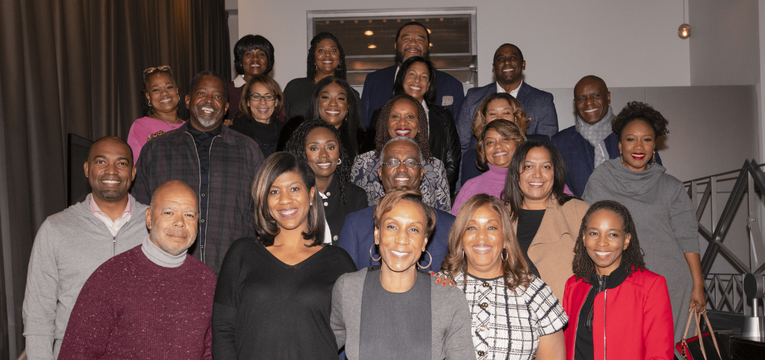A large group of diverse professionals poses together in an indoor setting, smiling warmly at the camera. They are dressed in business and business-casual attire, with a mix of colors and styles. The setting features modern lighting and a welcoming atmosphere, suggesting a networking or leadership event. The group stands closely together, reflecting a sense of camaraderie and community.