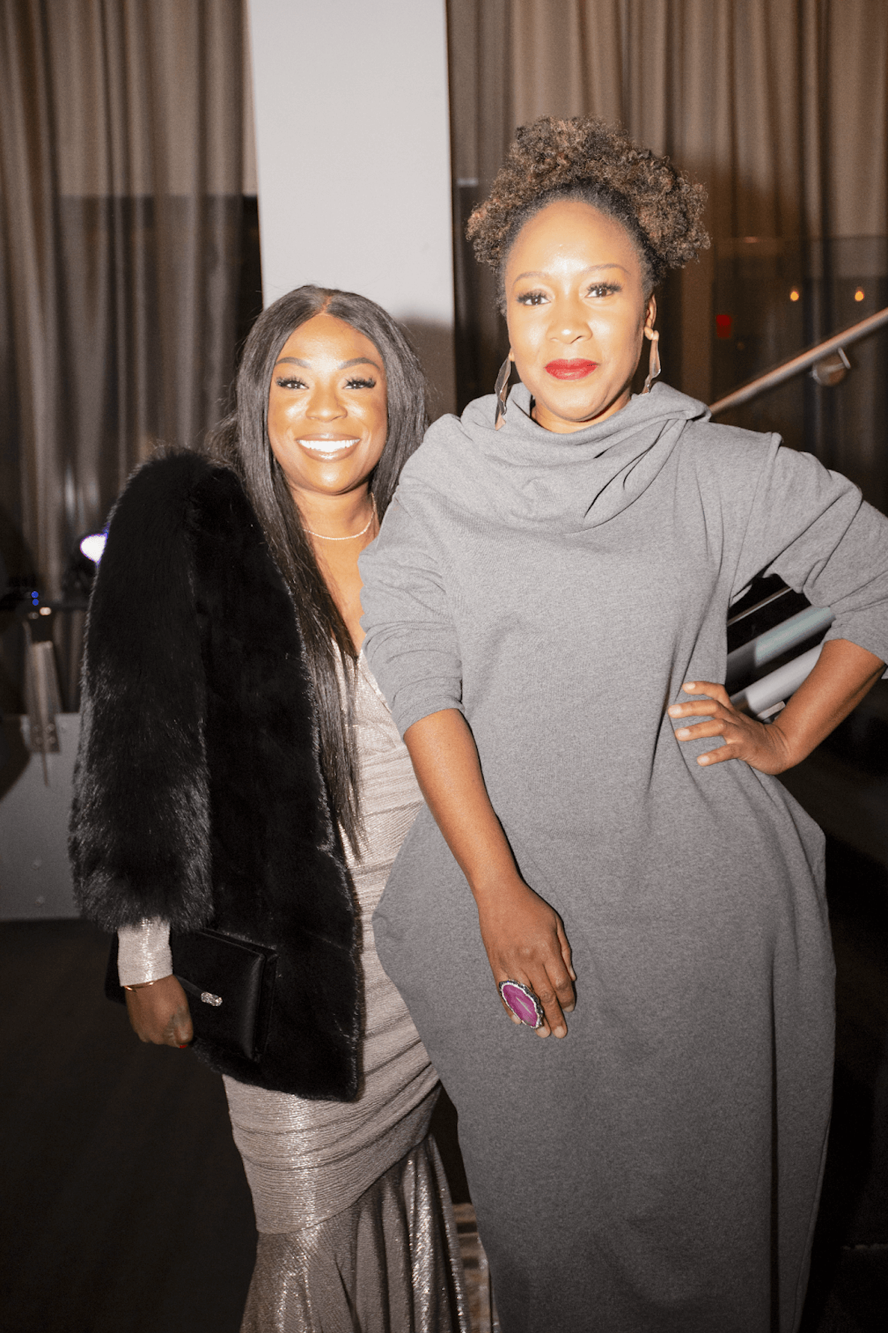 Two elegantly dressed women pose together, one in a glamorous silver gown with a fur shawl and the other in a cozy yet chic gray outfit, smiling warmly at the camera.