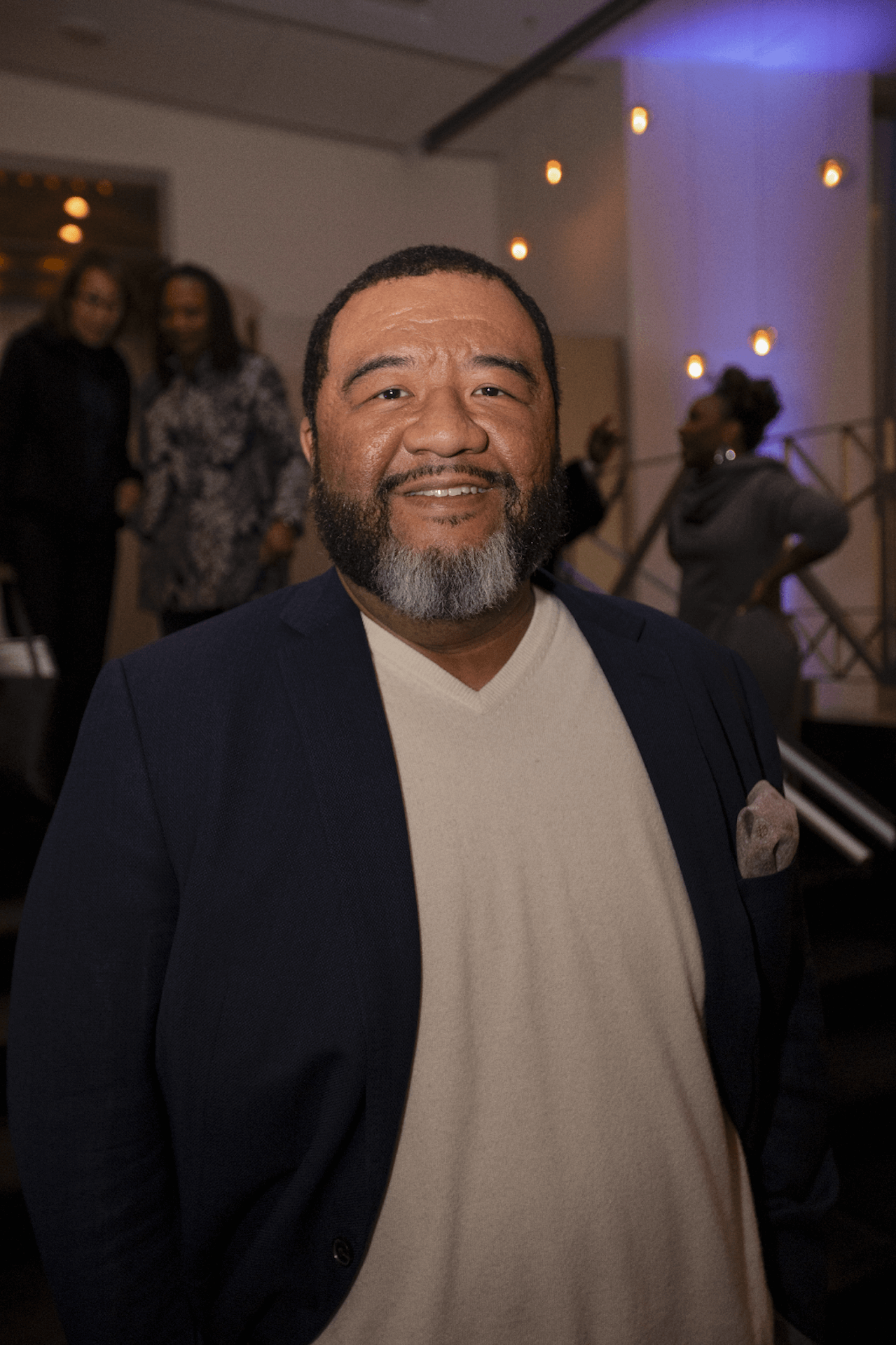 A man with a beard smiles warmly at the camera, dressed in a navy blazer and beige shirt. The background features a softly lit event space with people and decorative lights.