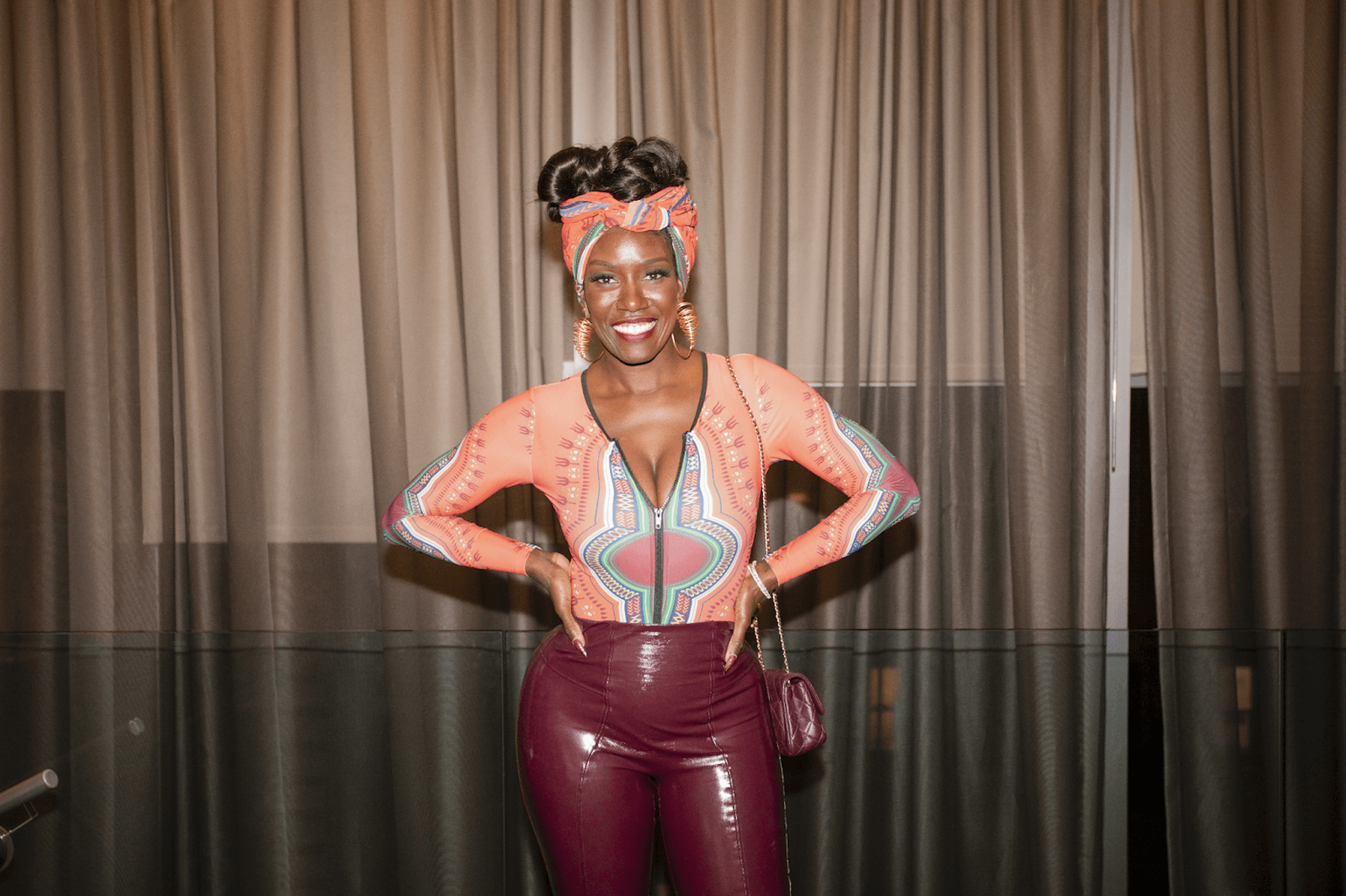 A vibrant individual wearing a bright orange top with intricate patterns, paired with burgundy pants and a matching headwrap, smiling confidently in front of drapes.