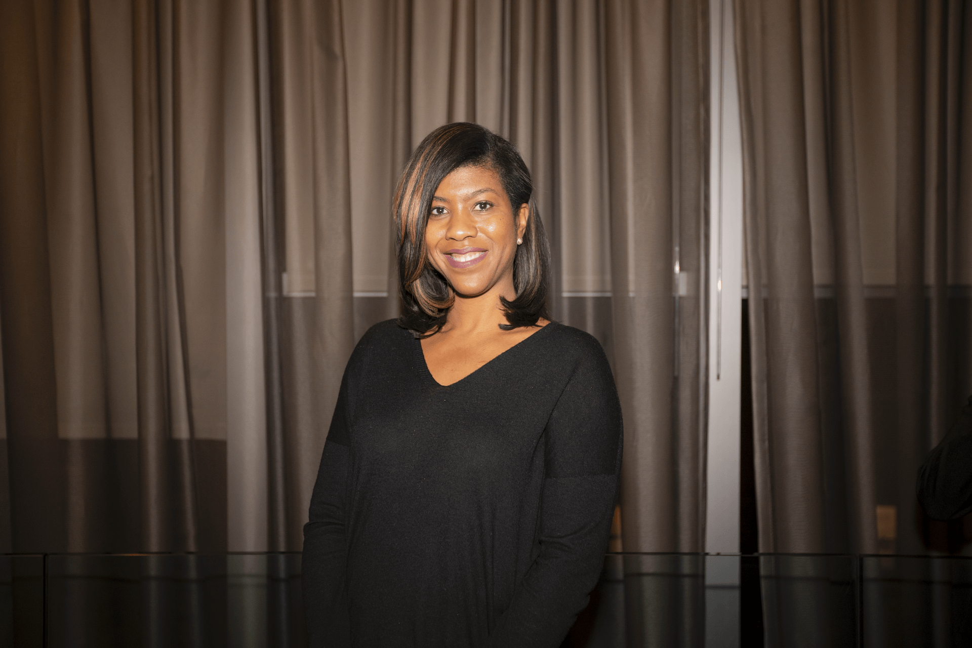 A woman in a simple black outfit, smiling softly while standing against a background of neutral drapes.