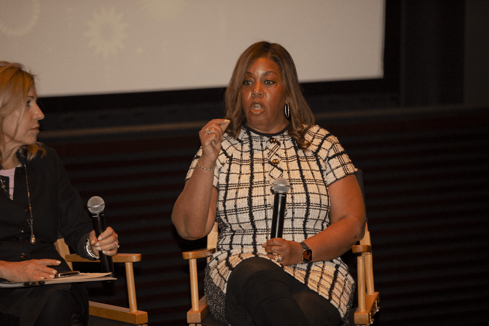 A panelist in a checkered dress passionately speaking into a microphone, with another panelist sitting beside her.