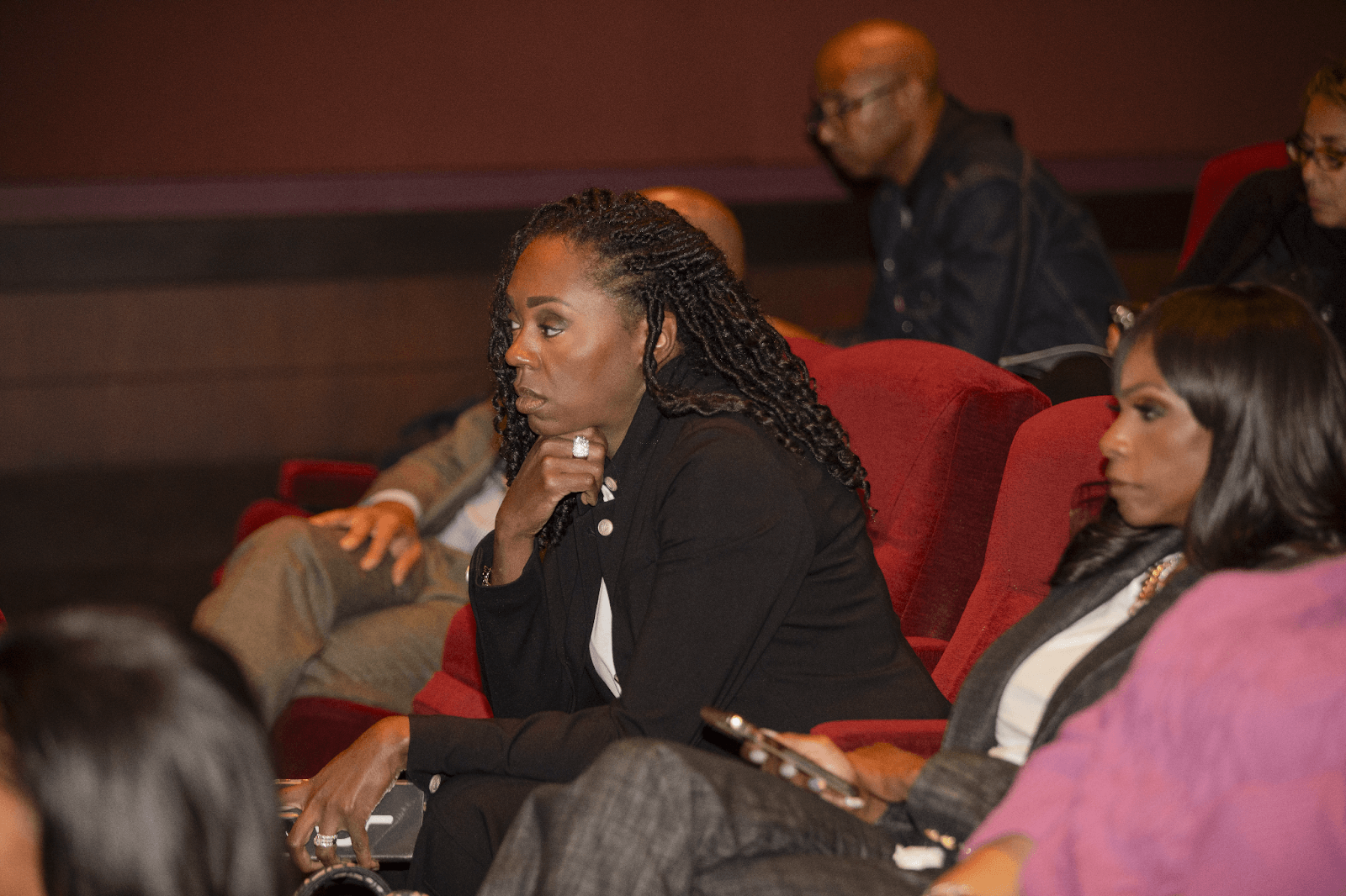 A woman in professional attire sits attentively in a red theater seat, appearing engaged in a discussion or presentation.